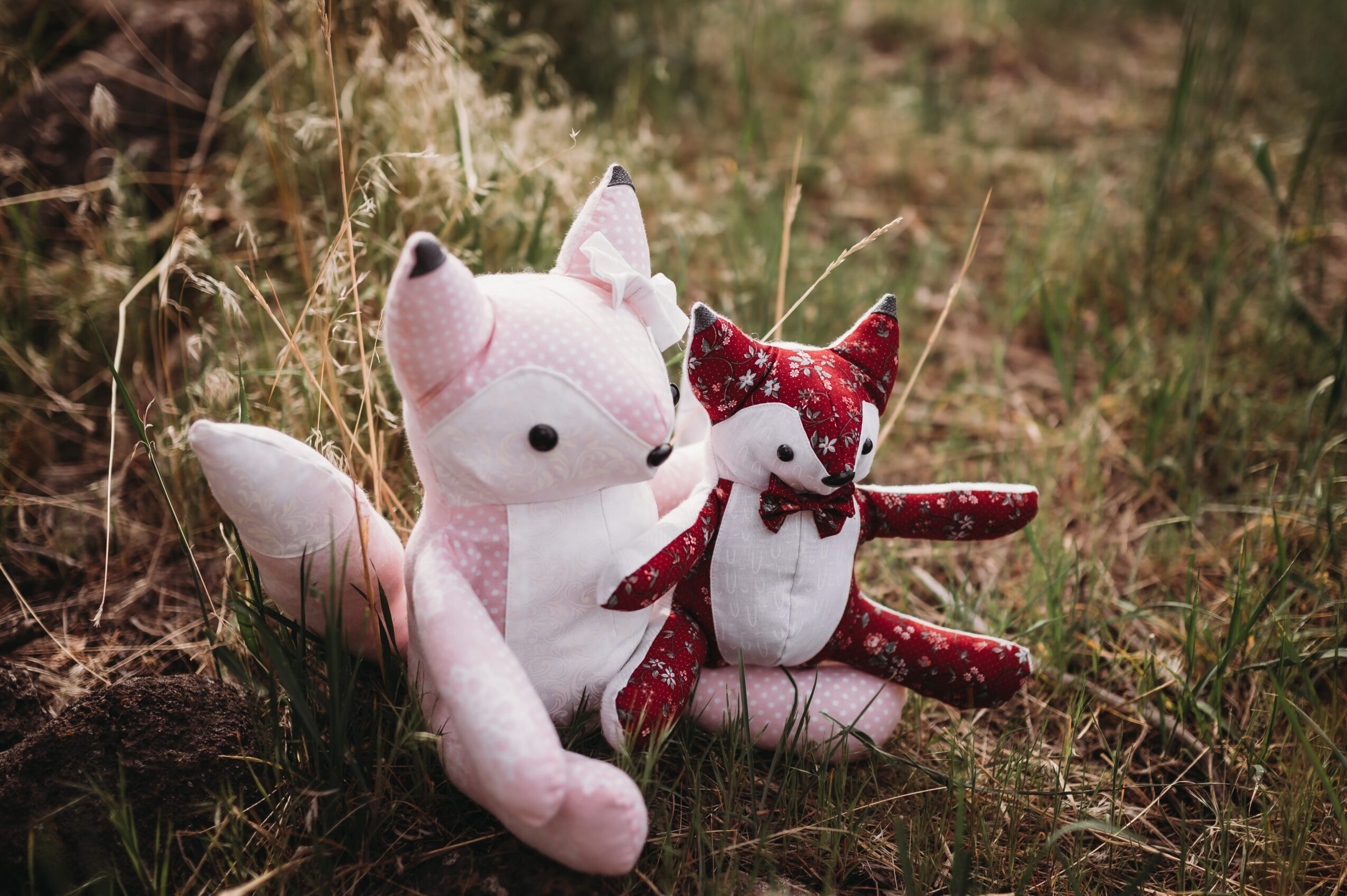 Two plush fox toys sit on grass with plants in the background, one larger pink and white, one smaller red and white.