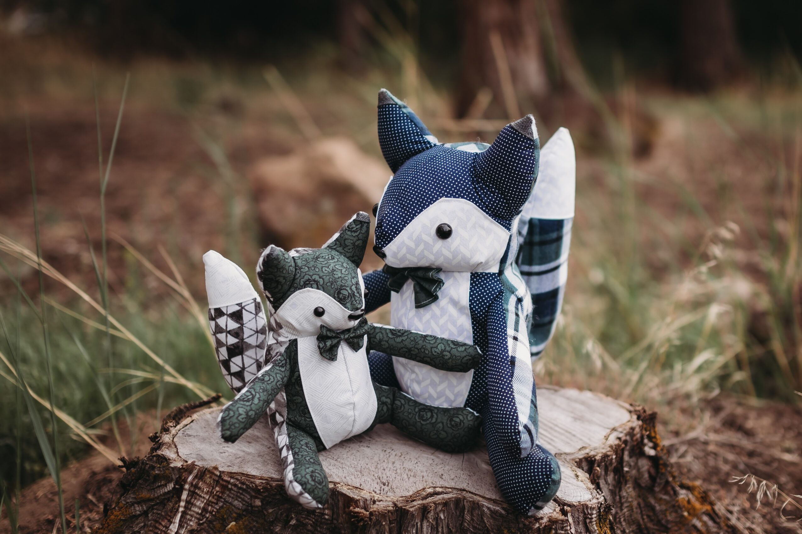 Two plush animal toys, a fox and a raccoon, sit on a tree stump outdoors with grass and plants in the background.