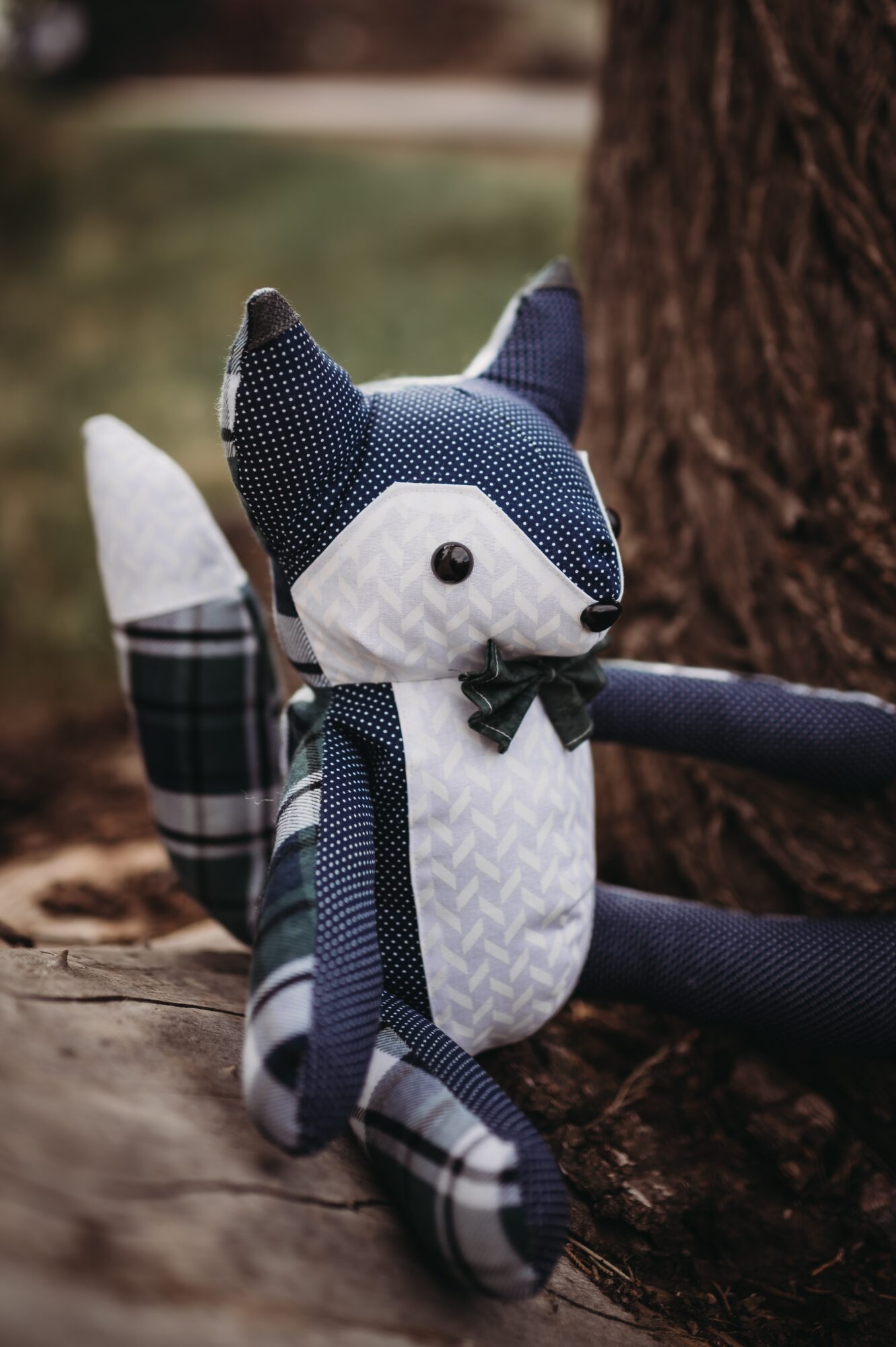 Plush fox toy with a bow tie, sitting outdoors near a tree trunk, with blurred background.