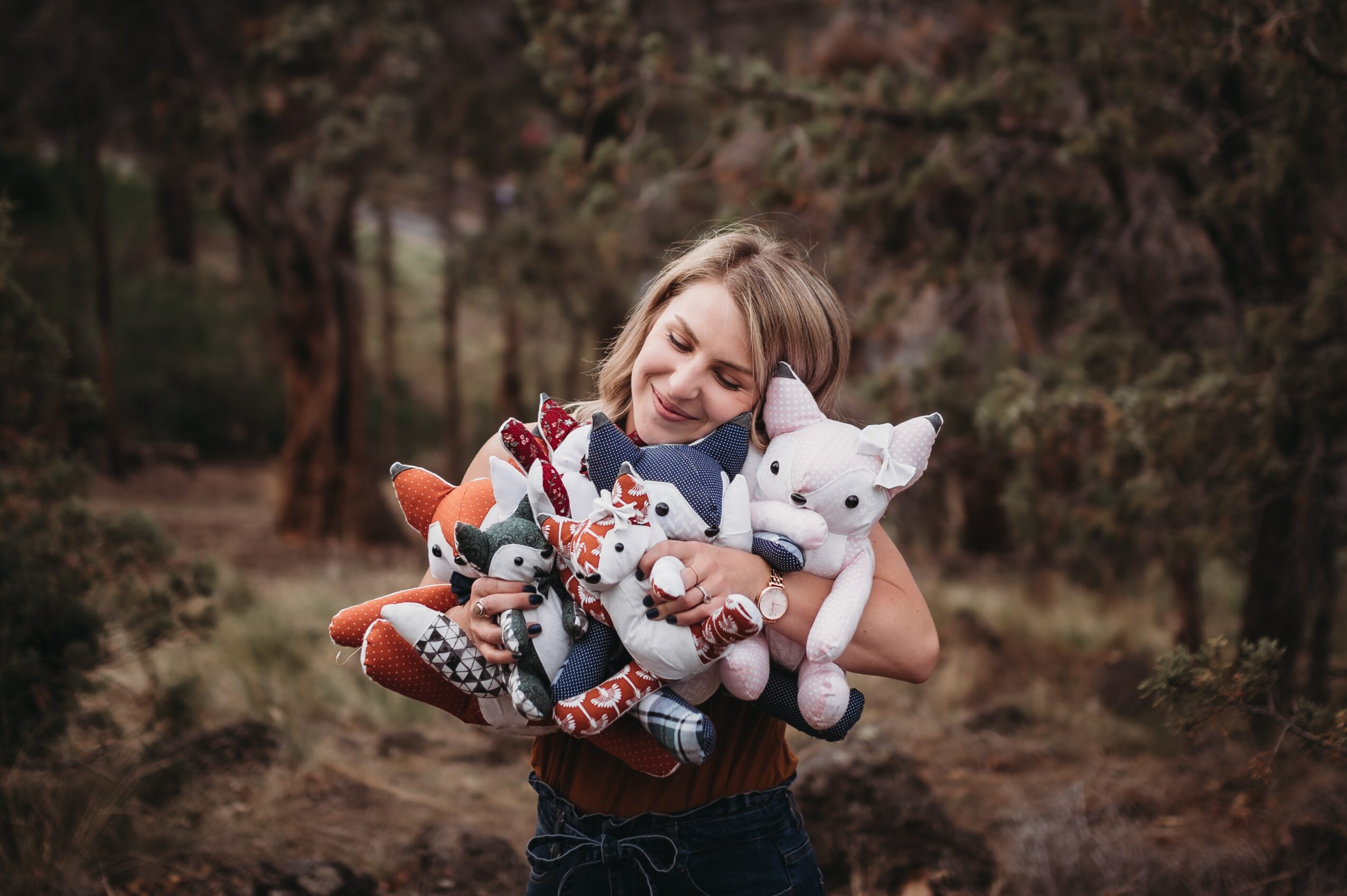 Woman hugging plush toys outdoors, smiling, surrounded by trees and nature.