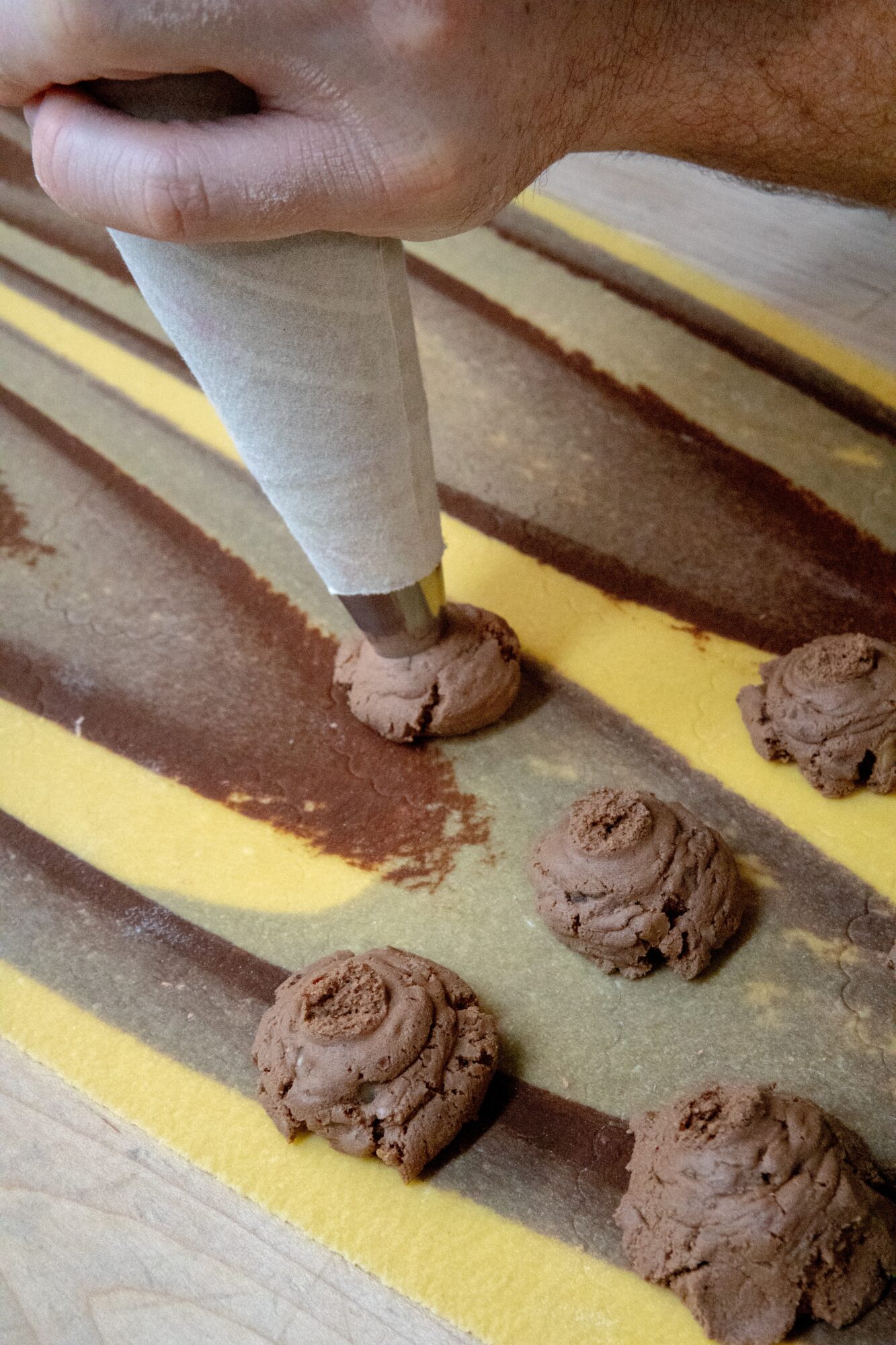 Person piping chocolate onto striped yellow and brown surface with a piping bag, chocolate cookies nearby.