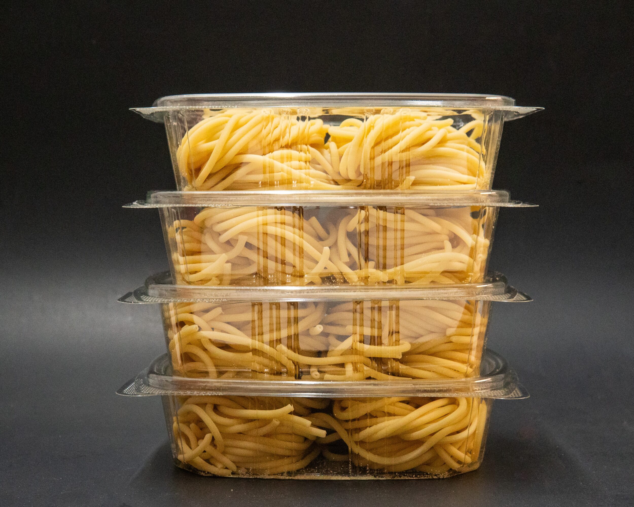 Four stacked clear plastic containers filled with cooked spaghetti against a black background.