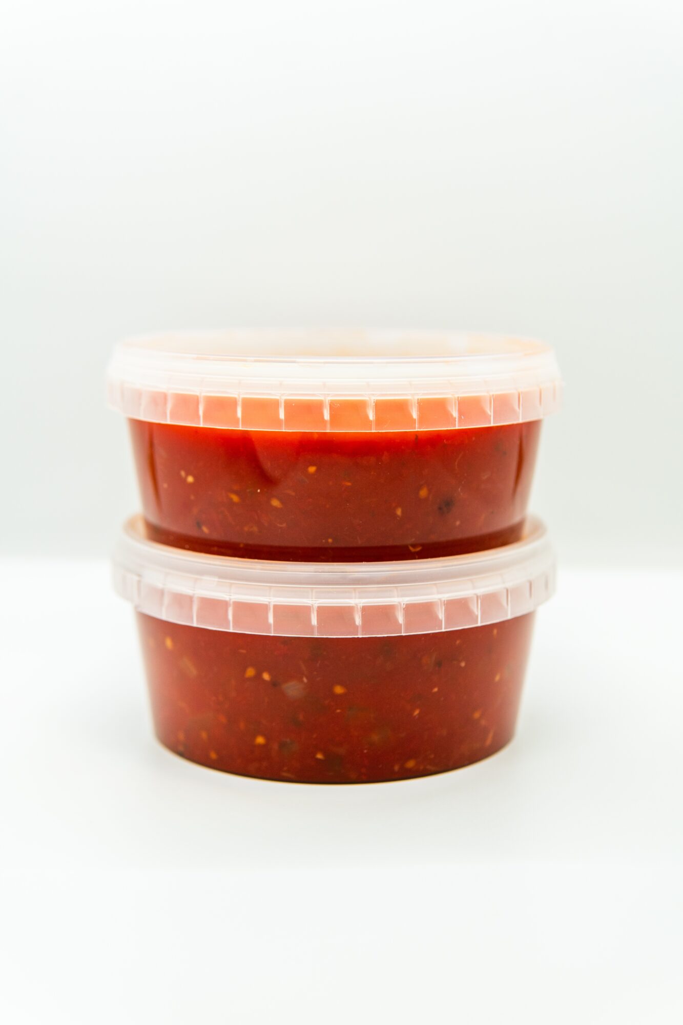 Two stacked clear plastic containers filled with red sauce or jam, with lids, on a white background.