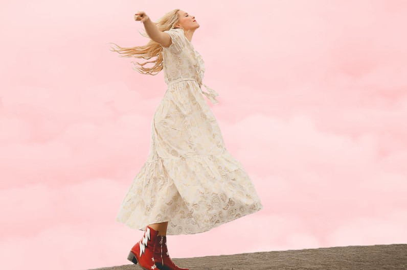 Woman in a long, light-colored dress with arms raised, standing outdoors against a pink sky background.