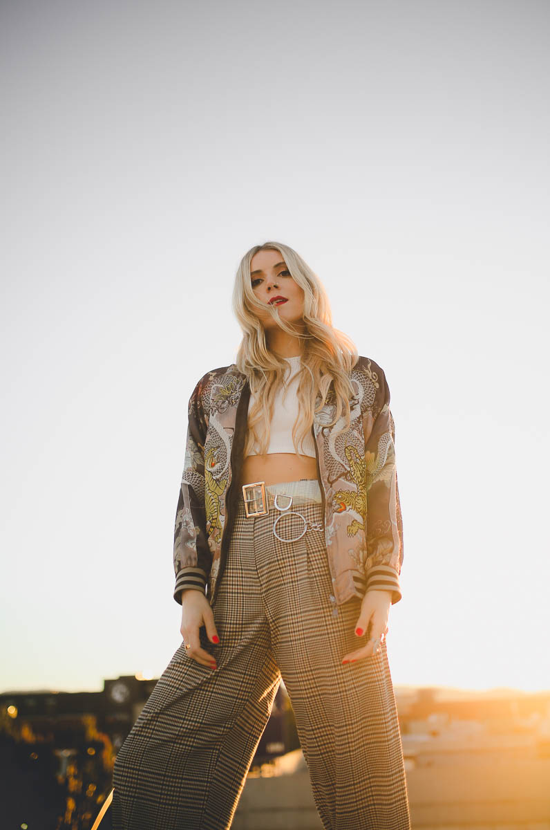 Young woman with long blonde hair standing outdoors during sunset, wearing a patterned jacket and wide-leg pants, looking at the camera.