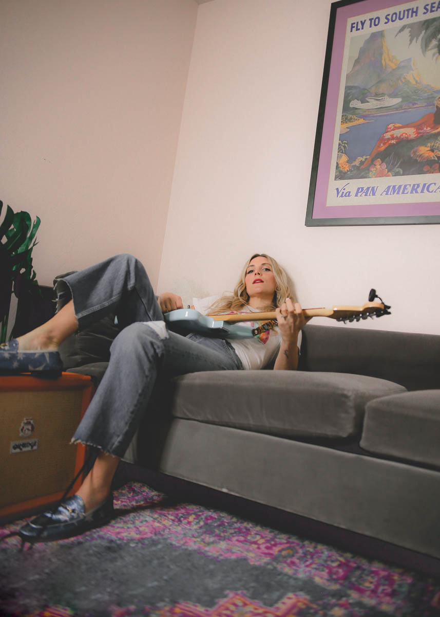 Person lying on a sofa holding a guitar, with a poster on the wall behind them.