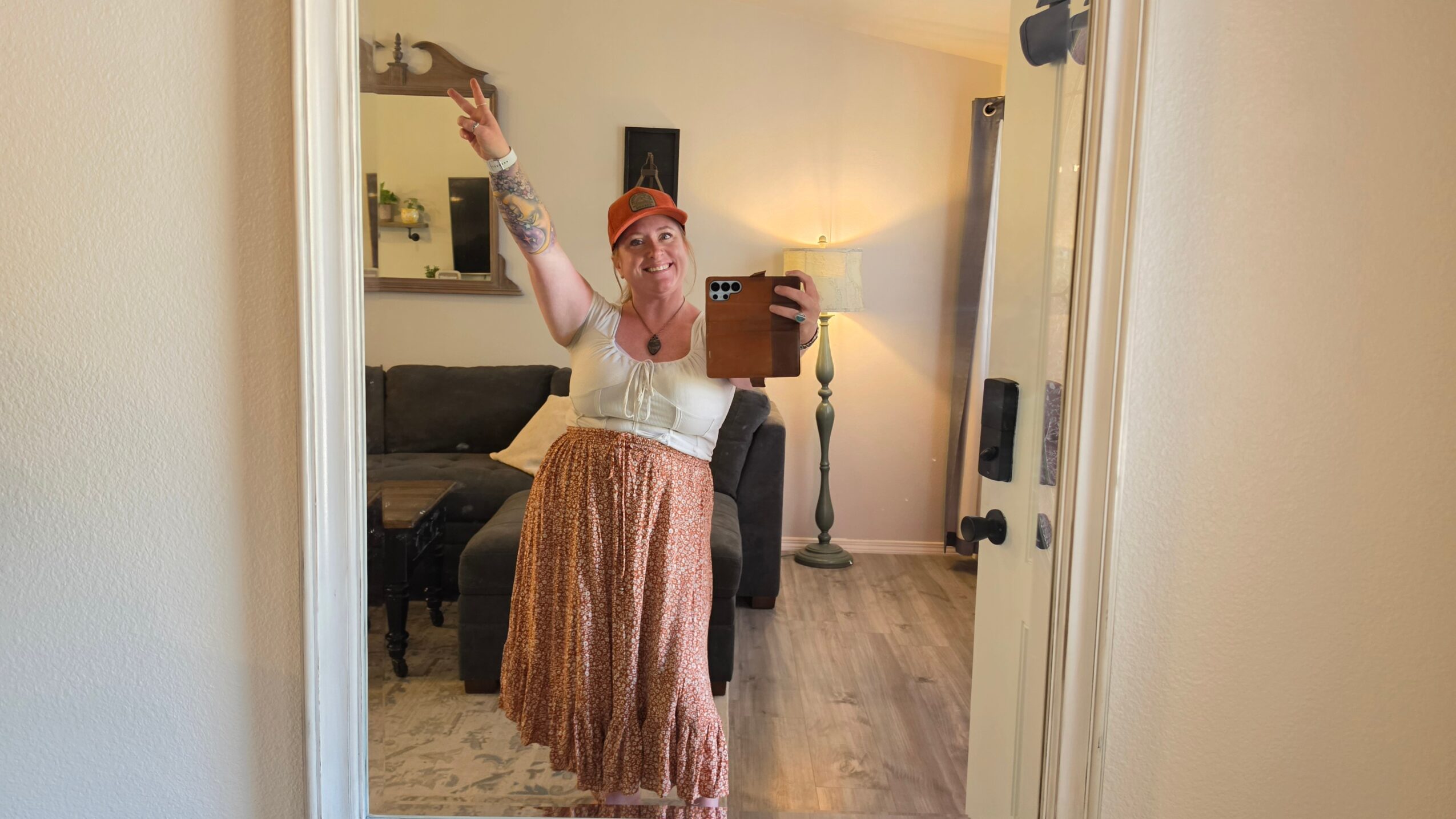 Person taking a mirror selfie in a room, wearing a pink headscarf, white top, and patterned skirt, holding a phone.