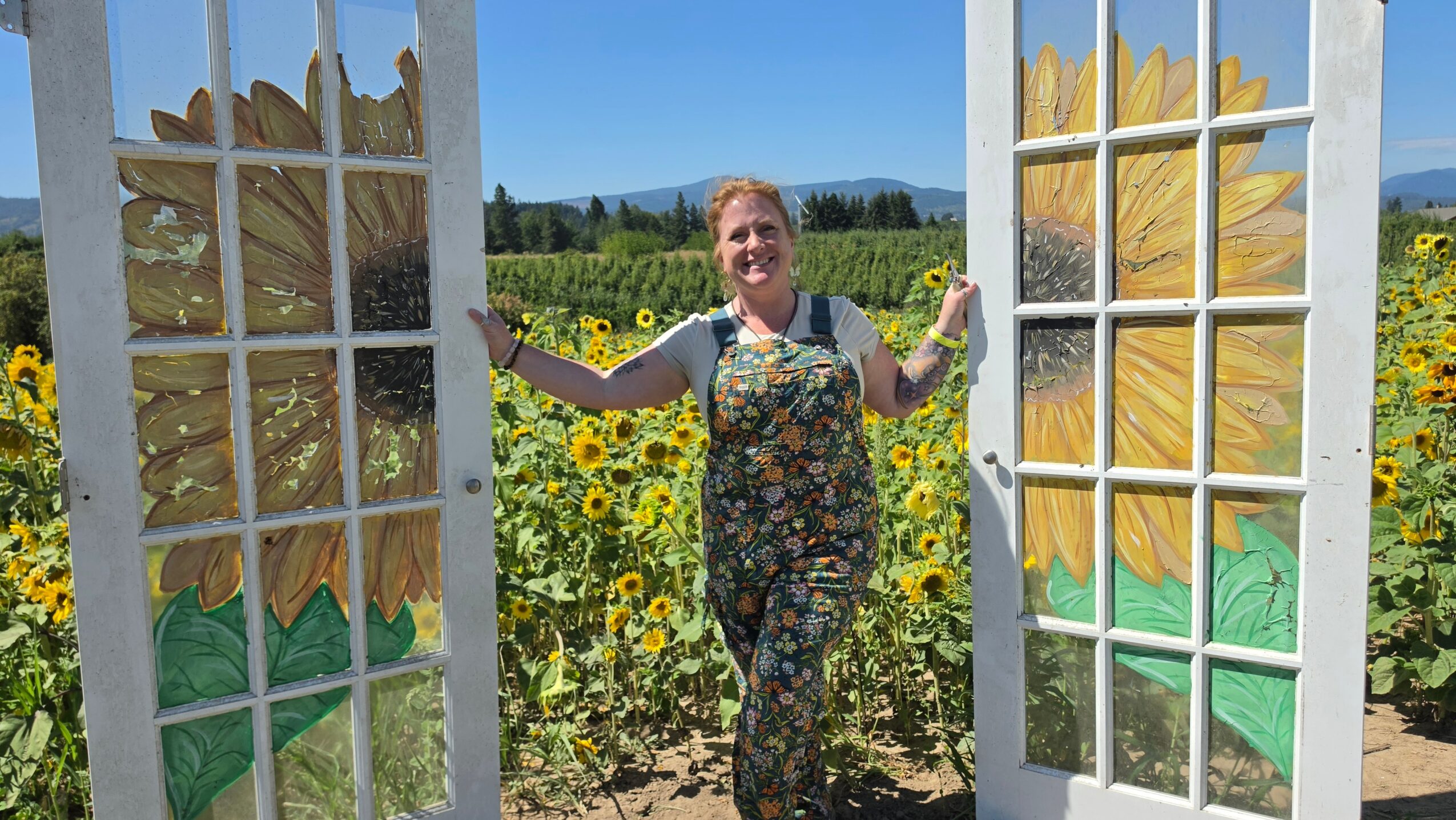 Woman standing between painted sunflower doors in a sunflower field, smiling, with mountains in the background.