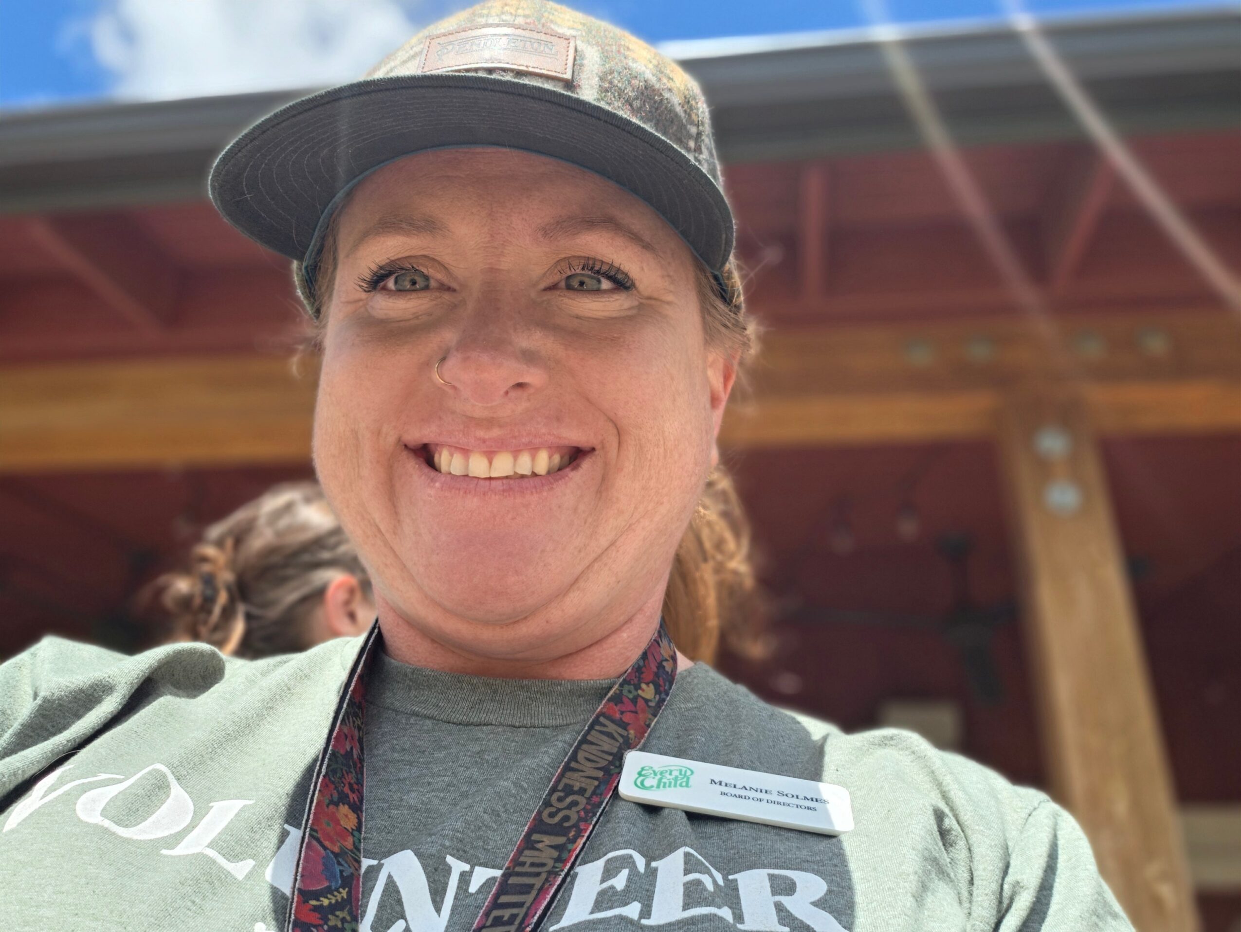 Smiling woman wearing a cap and a name tag outdoors, with a wooden structure in the background.