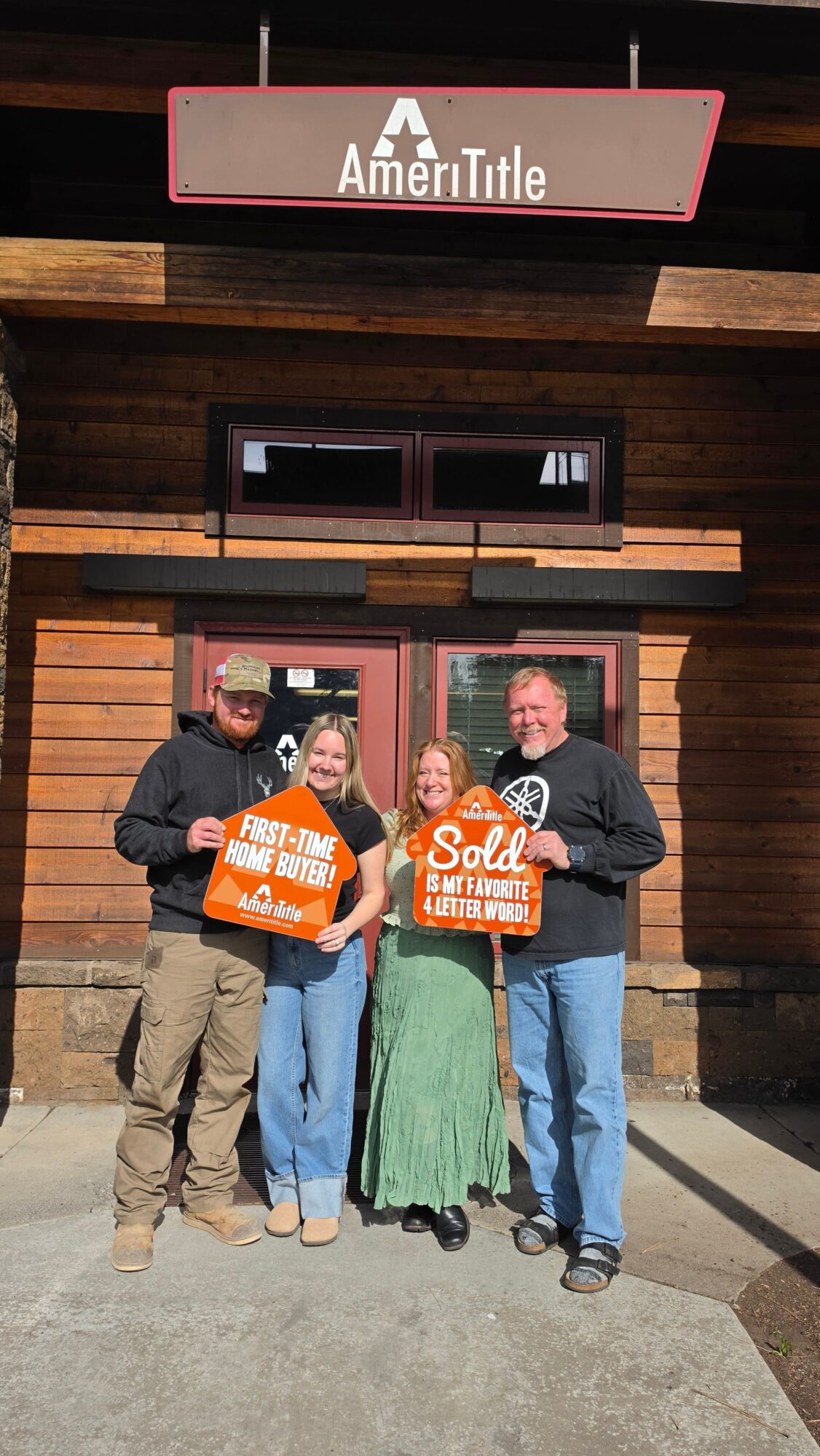 Three people standing outside a building holding orange signs, with a wooden wall and window behind them.