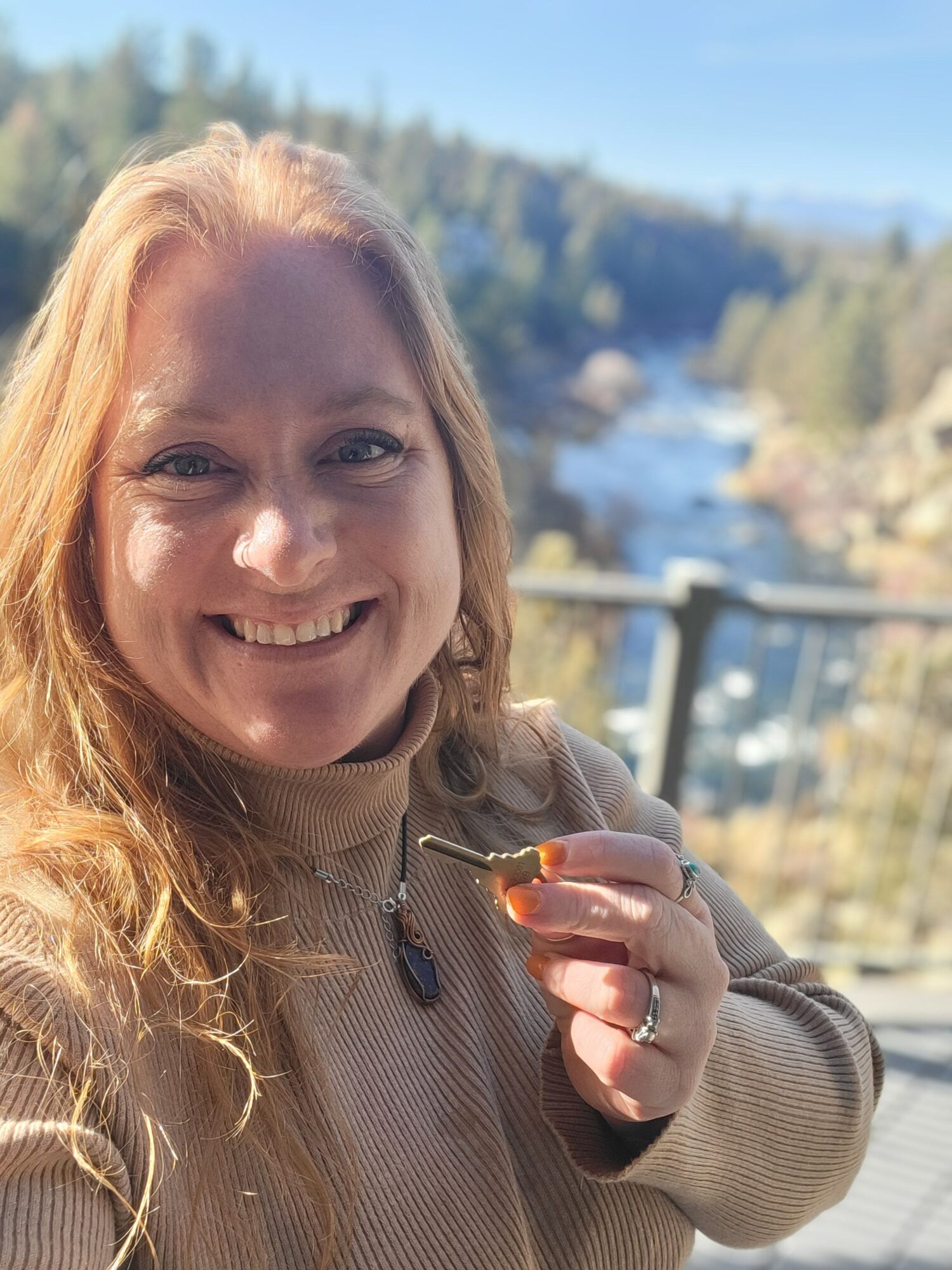 Woman smiling outdoors holding a small object, with trees and water in the background.
