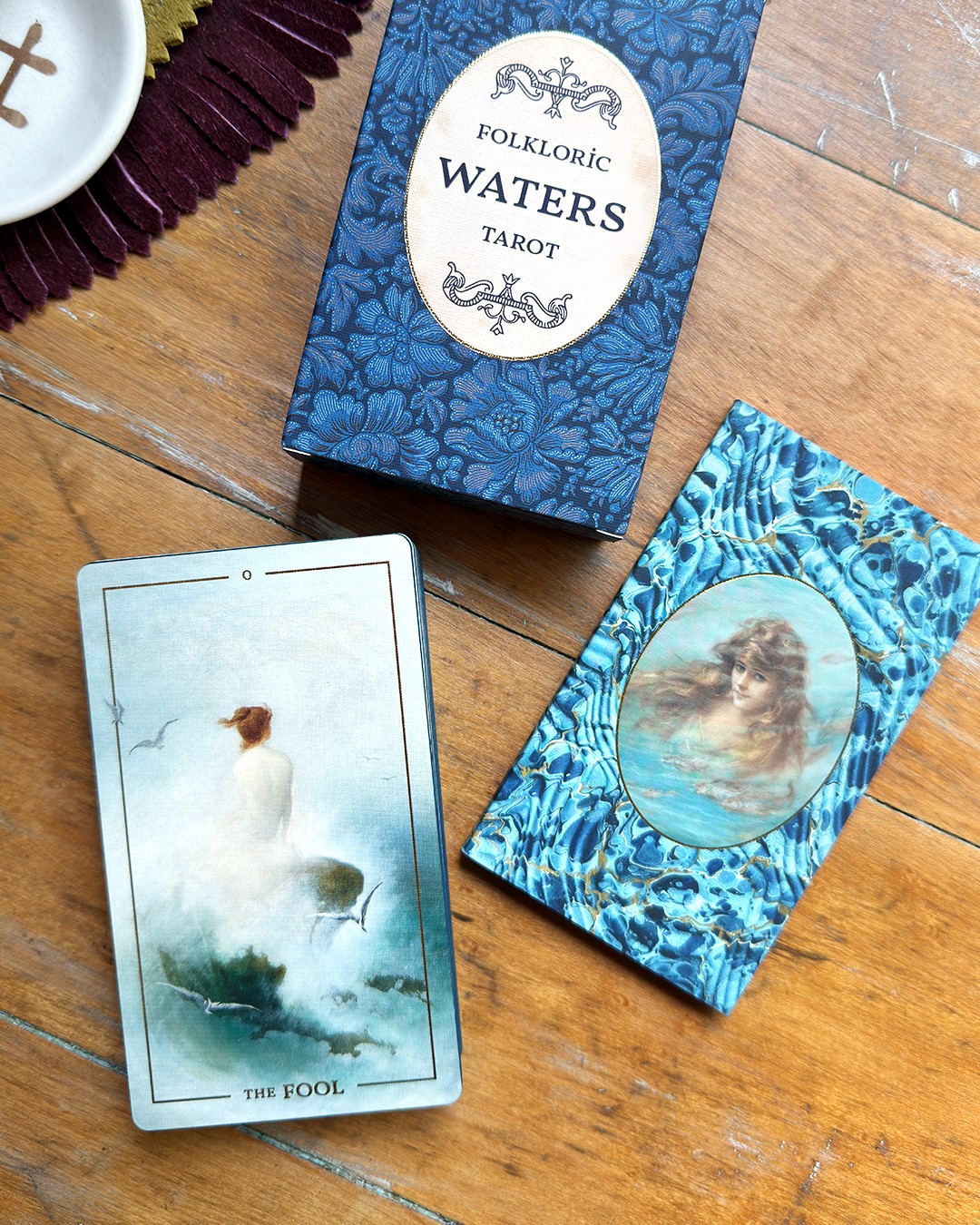 Water tarot card with a woman in a white dress sitting by a pool, surrounded by mist and trees, on a wooden surface.