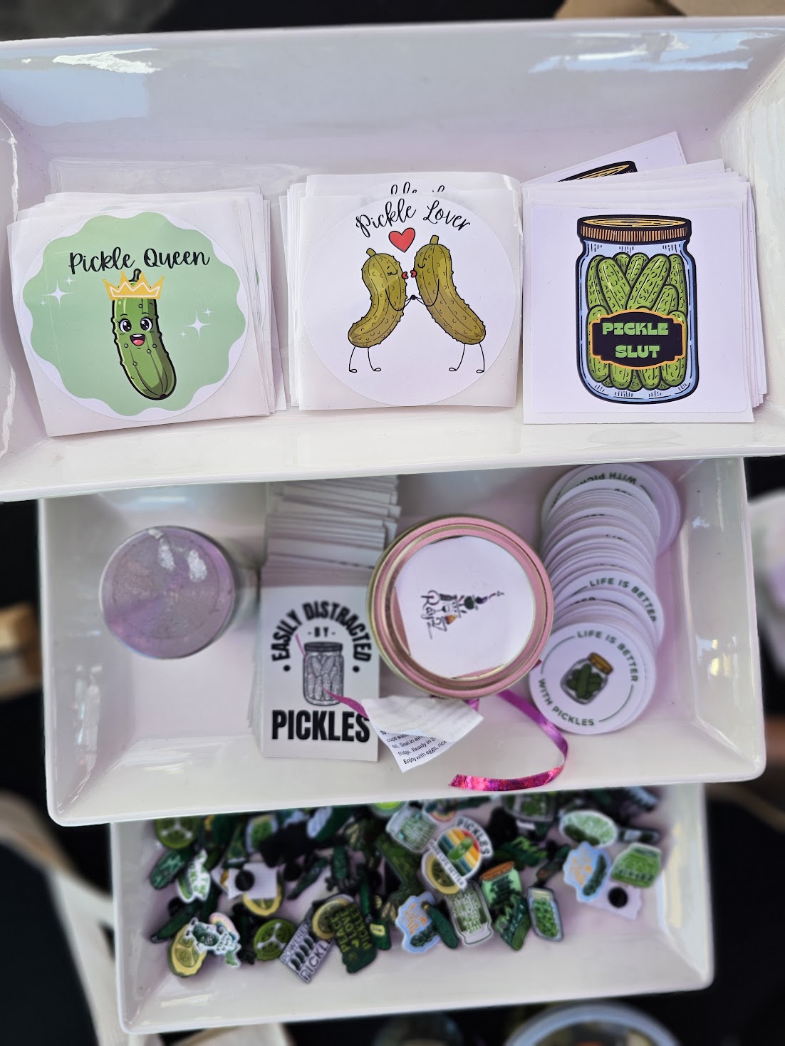 Three small boxes with pickle-themed illustrations on a white shelf, containing jars and accessories, with a lower shelf holding more items.