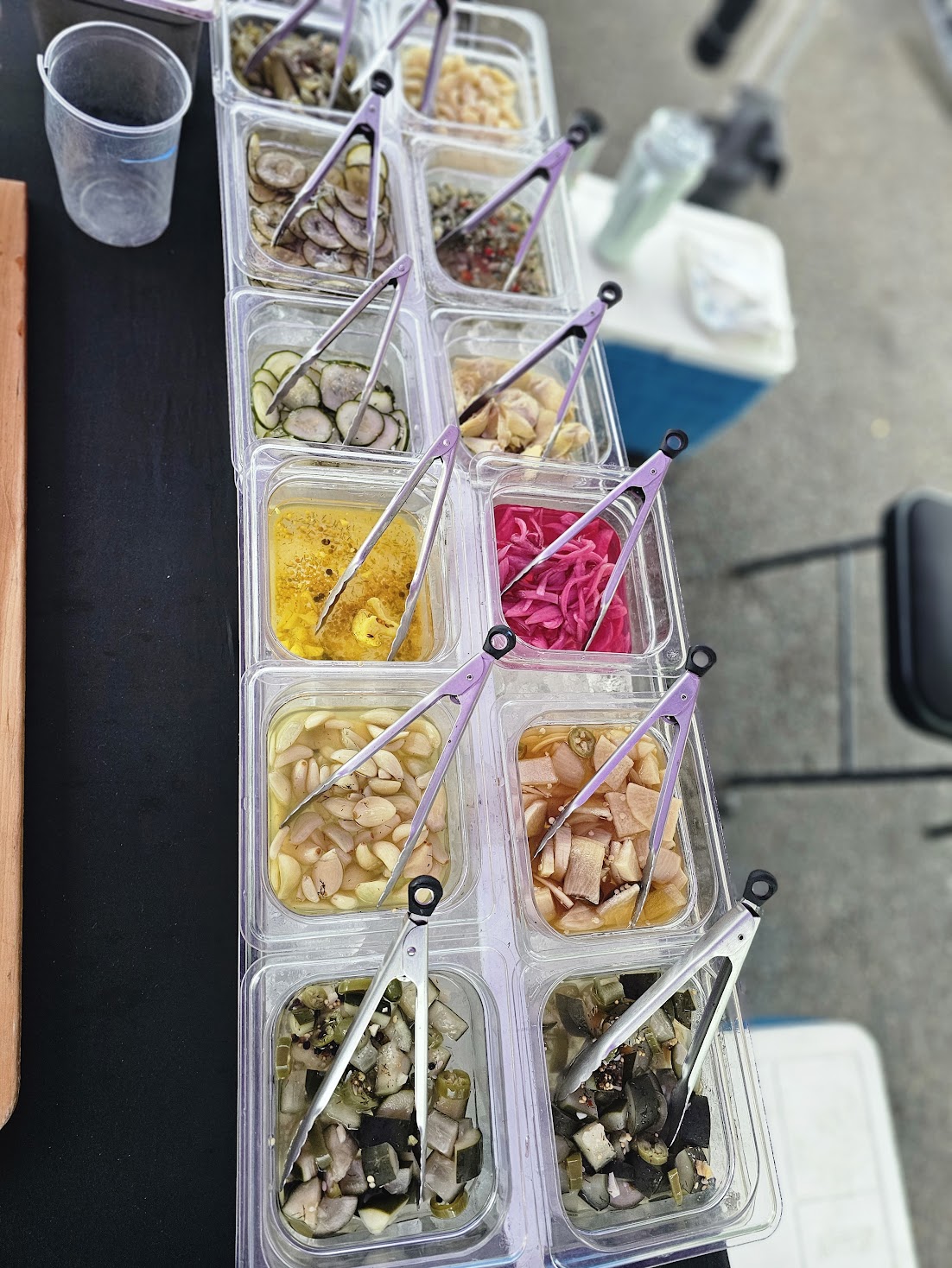 Multiple small containers with colorful toppings and white spoons on a black surface, with a plastic cup and other items nearby.