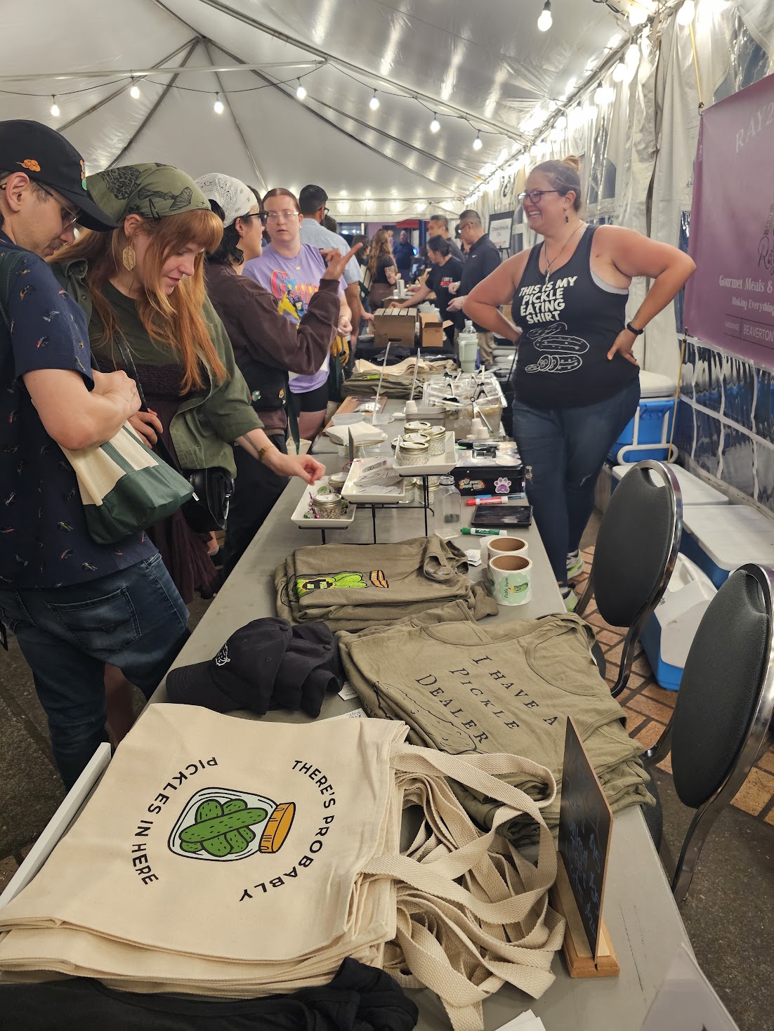 People gather around a table with bags, food, and items inside a large tent with string lights, engaging in an event.