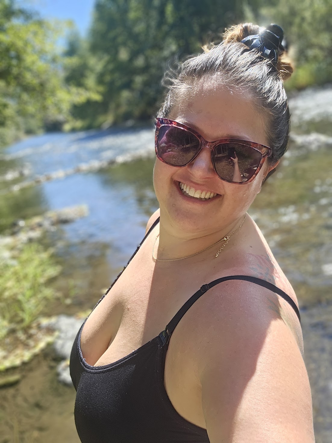 Smiling woman wearing sunglasses outdoors near a river with trees in the background.
