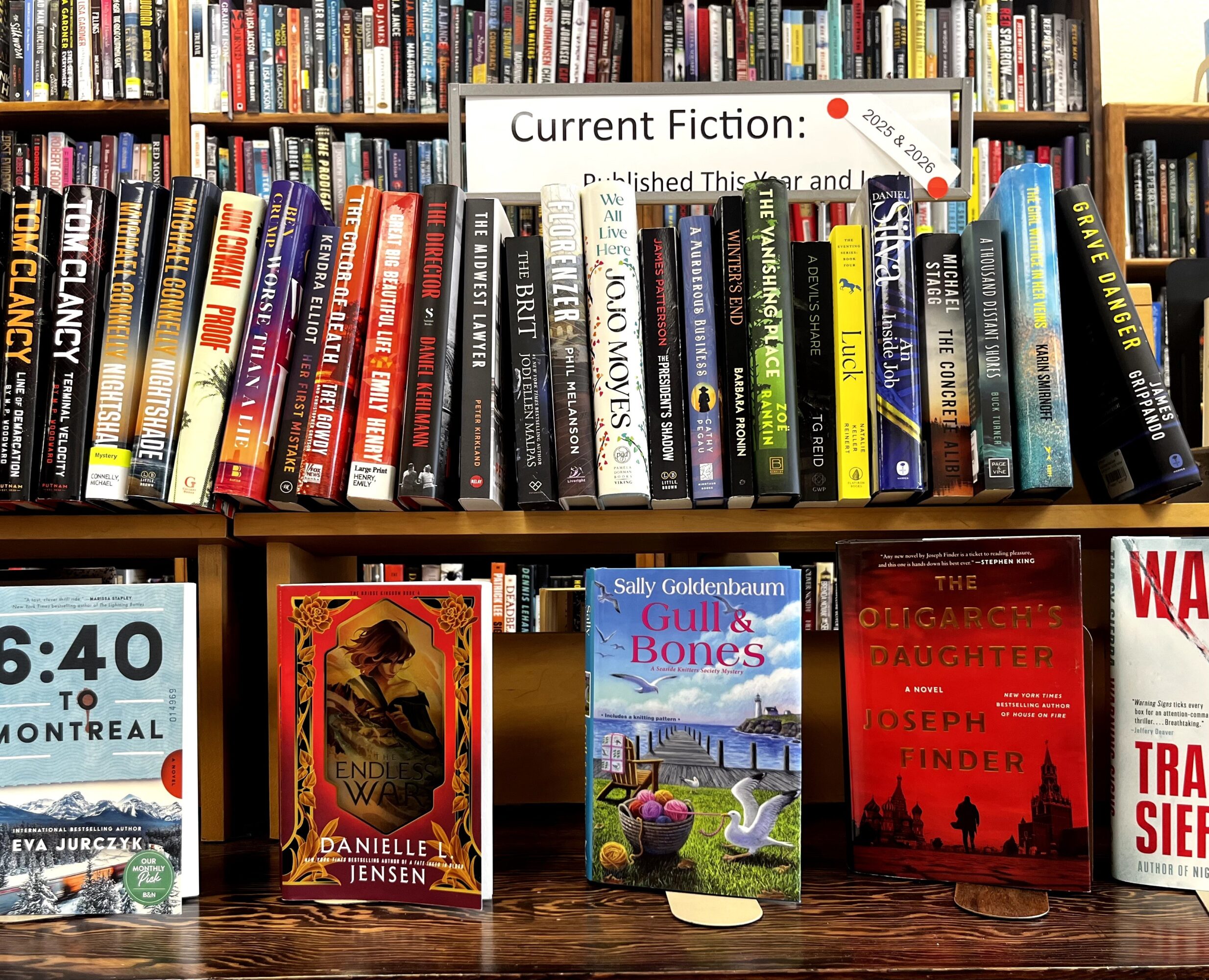 Books on a shelf and table, including fiction and non-fiction titles, with a sign about current fiction in the background.