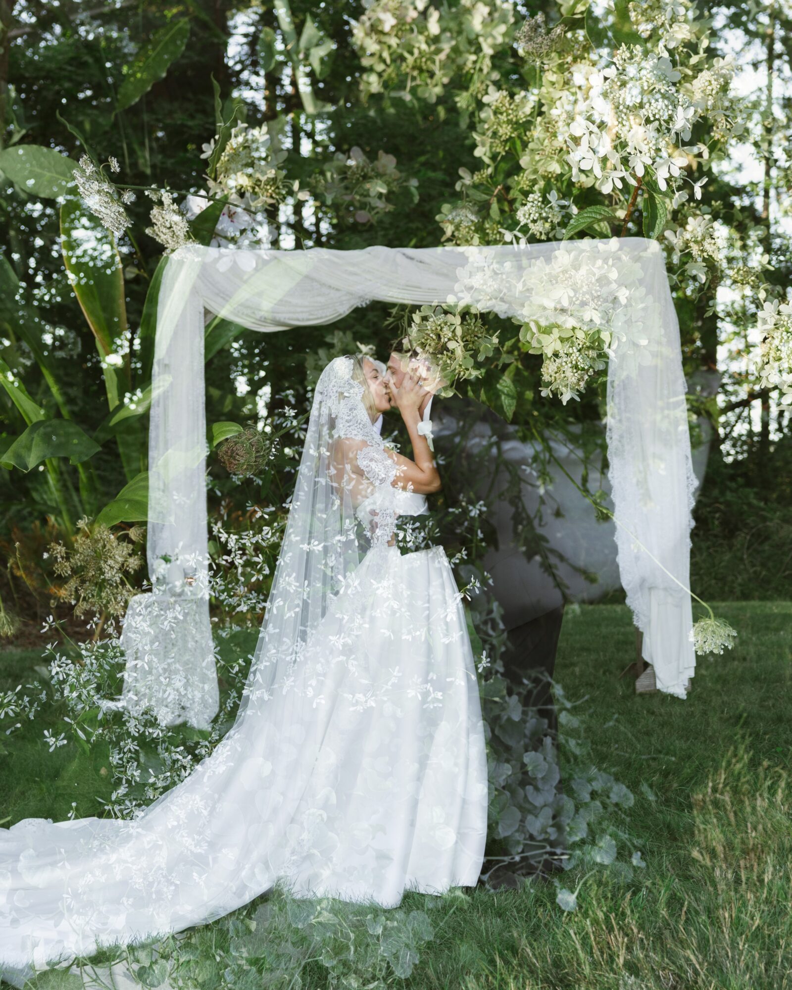 Bride in a white wedding dress and veil standing outdoors under a floral arch, surrounded by greenery.