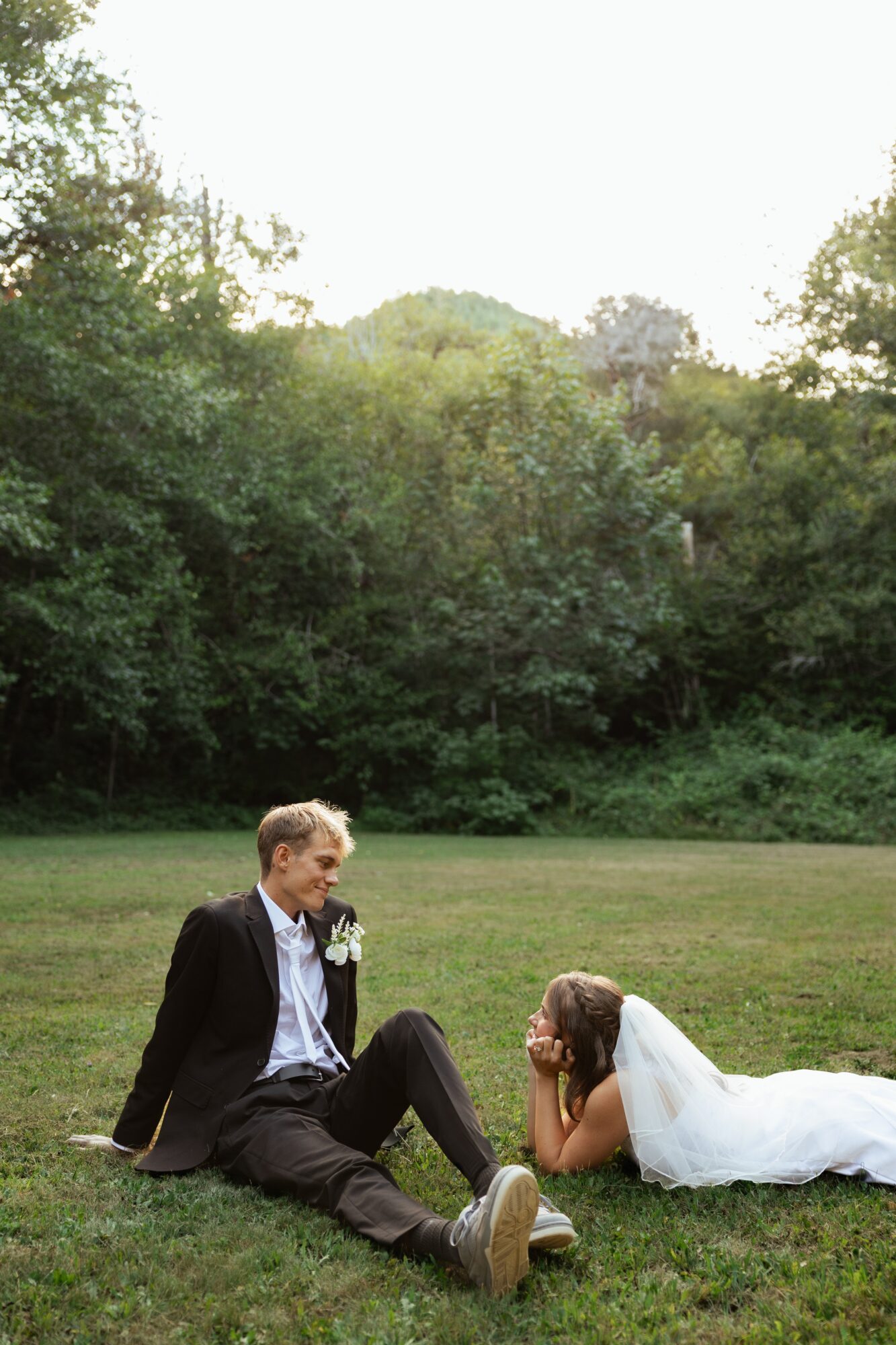 A man in a suit and a woman in a wedding dress sit on grass outdoors, with trees and sky in the background.