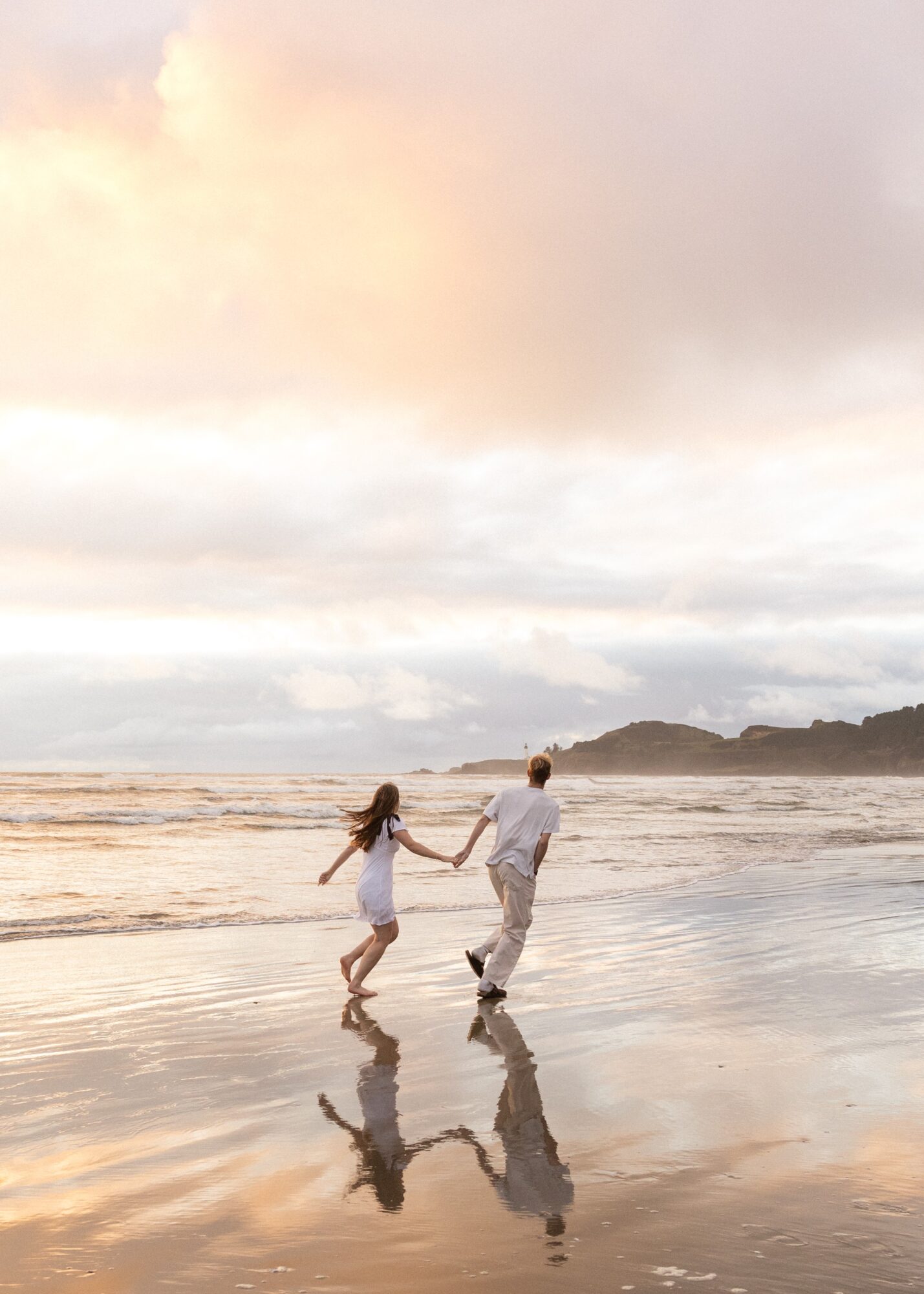 Two people holding hands walk along the beach with reflections in shallow water, cloudy sky, and distant landmass.