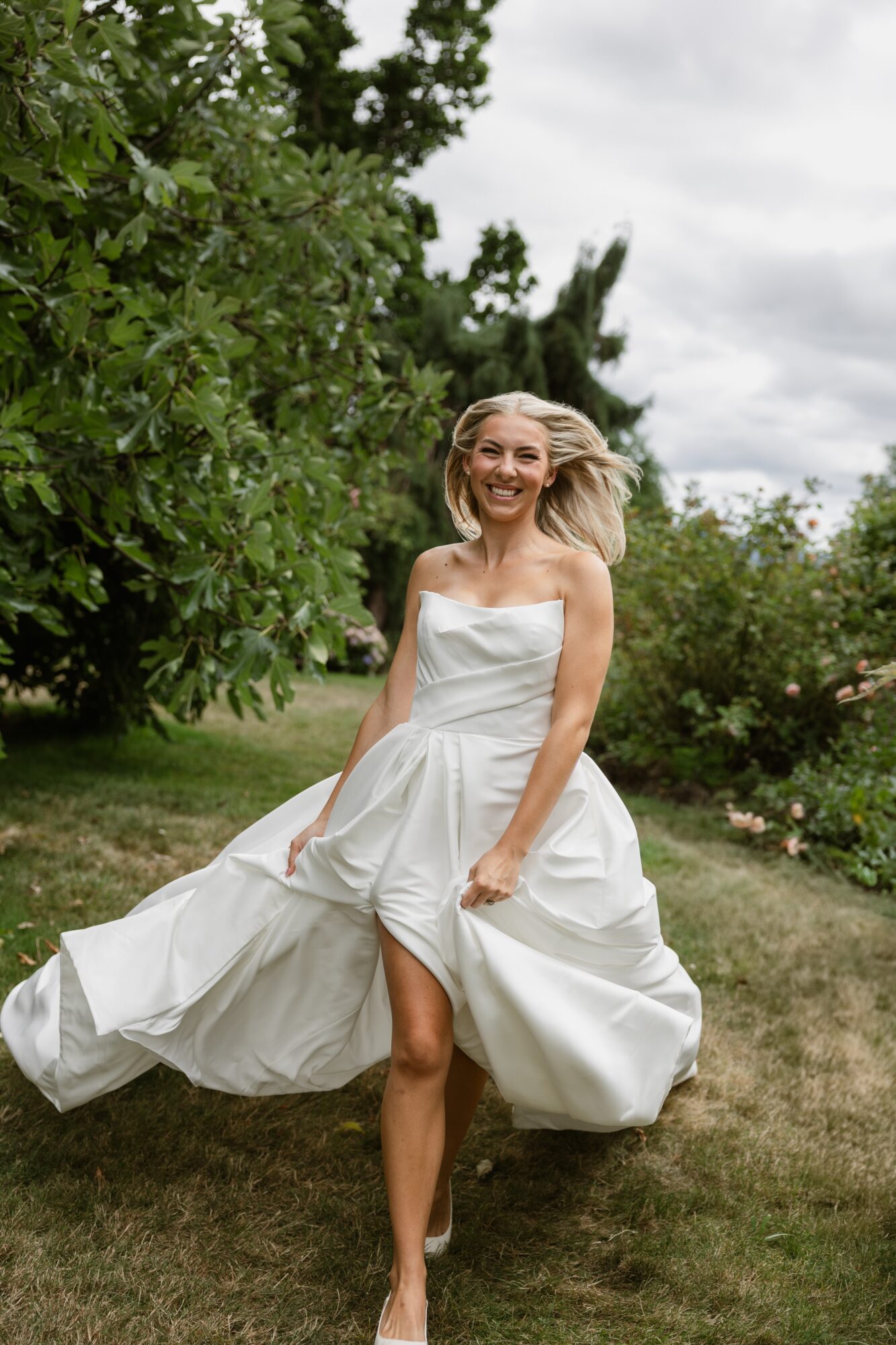 Woman in a white dress running outdoors, smiling, with trees and cloudy sky in background.