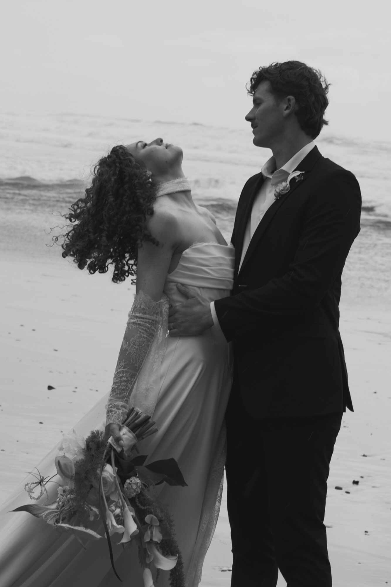 A couple stands on a beach, the woman in a dress holding a bouquet, the man in a suit, facing each other.