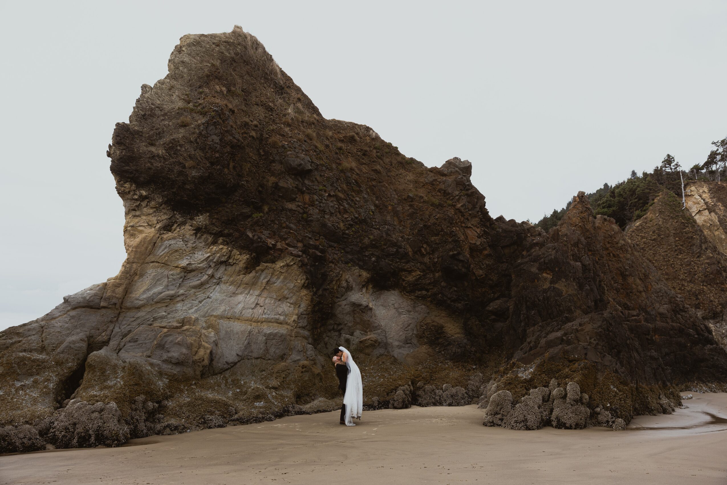 Person in white standing on sandy beach near large rock formation with overhanging cliff, cloudy sky background.