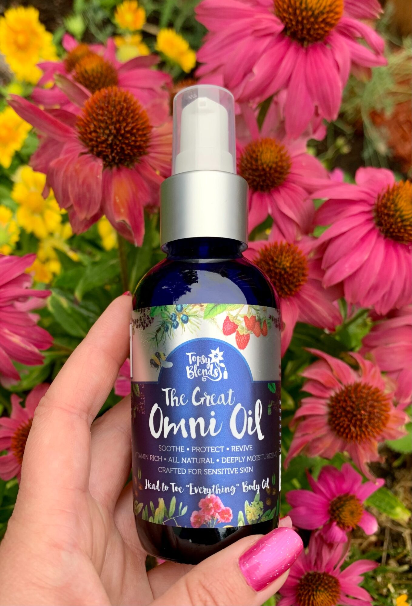 Hand holding a dark blue bottle of The Body Shop's Omni Oil with a white pump, surrounded by pink and yellow flowers.