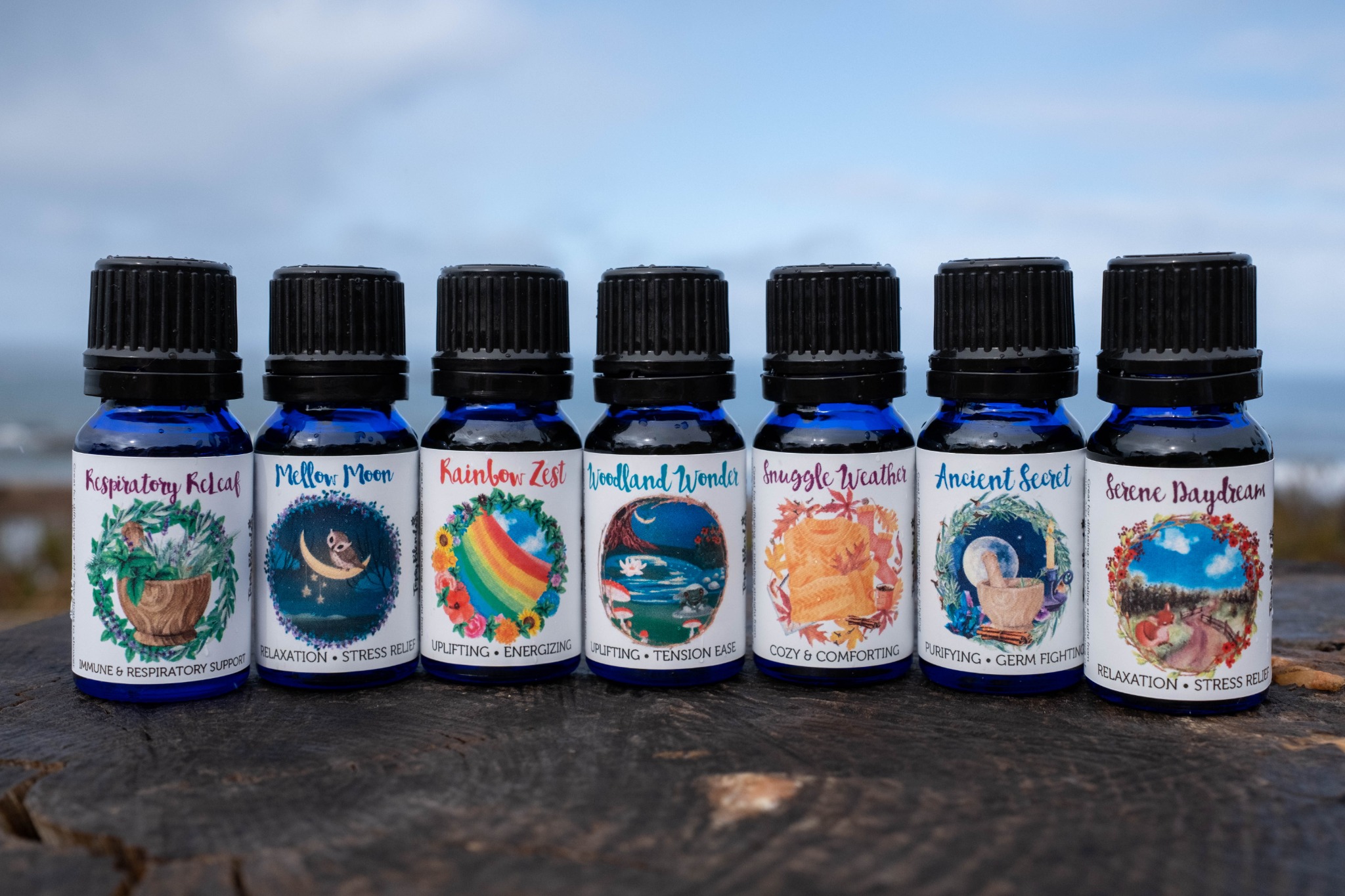 Seven small bottles with colorful labels arranged in a row on a surface outdoors, sky in background.