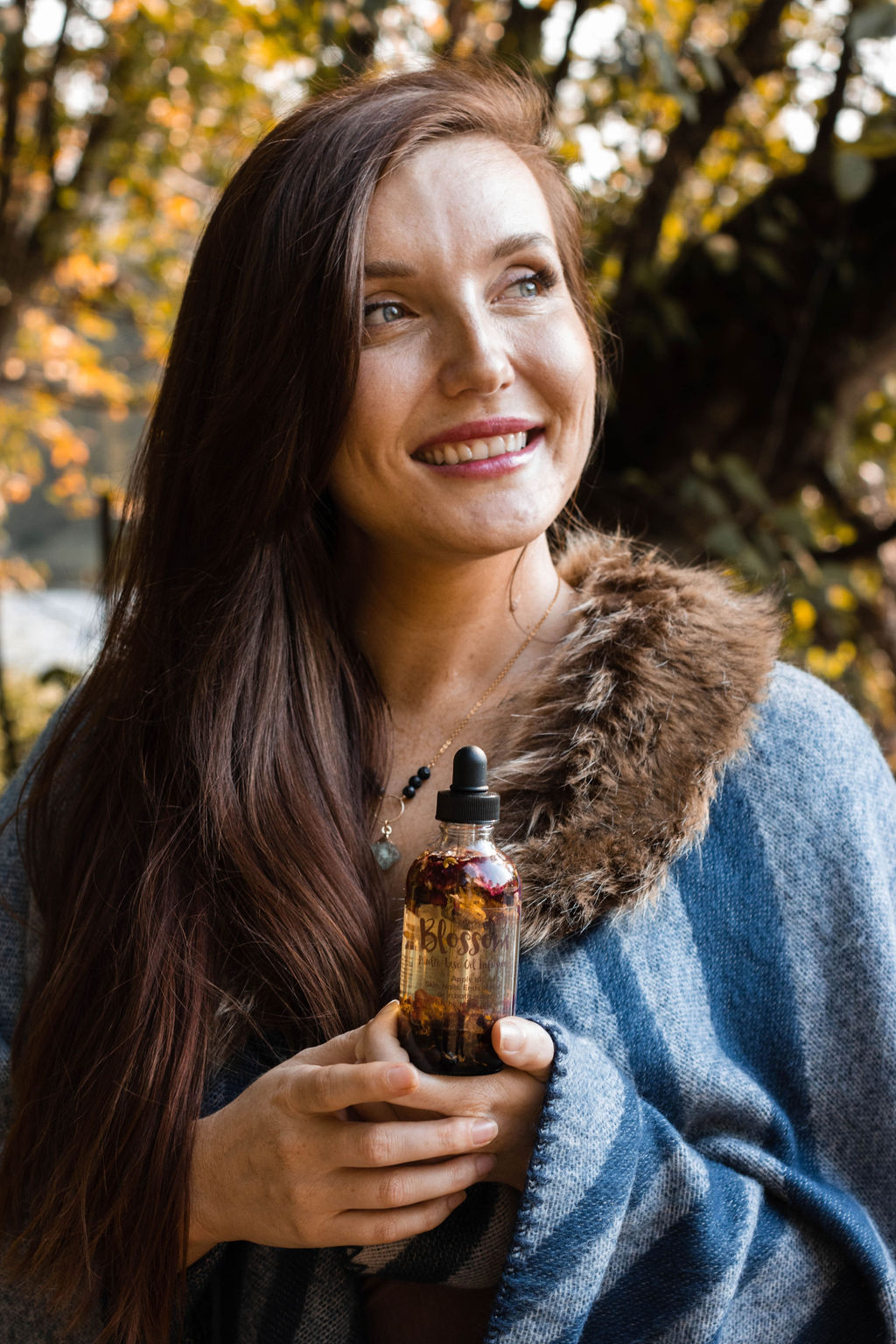 Woman with long dark hair smiling outdoors holding a small bottle, surrounded by autumn trees.