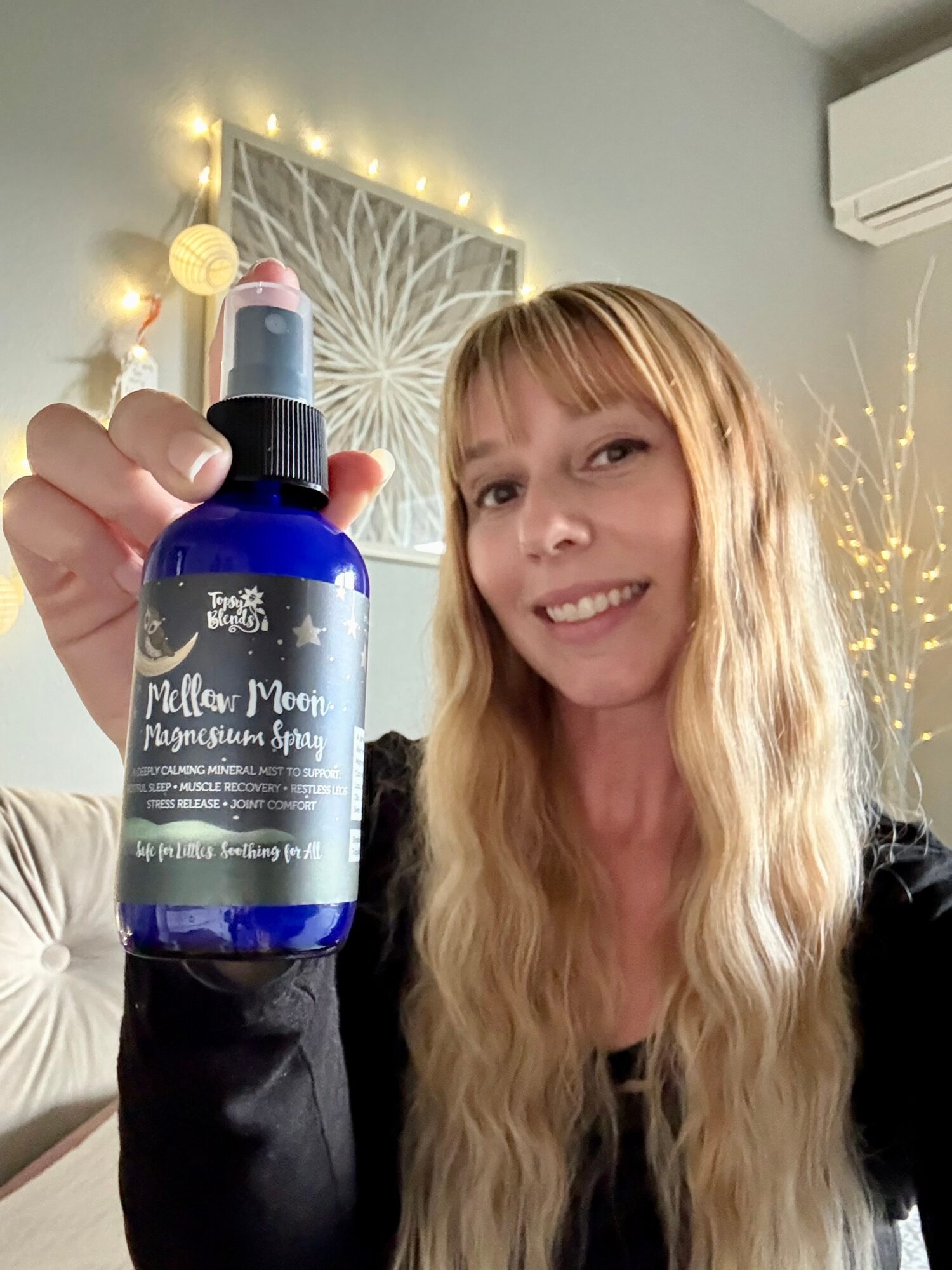 Woman with long blonde hair smiling, holding a blue spray bottle labeled 'Pollen Mace' in a decorated room.