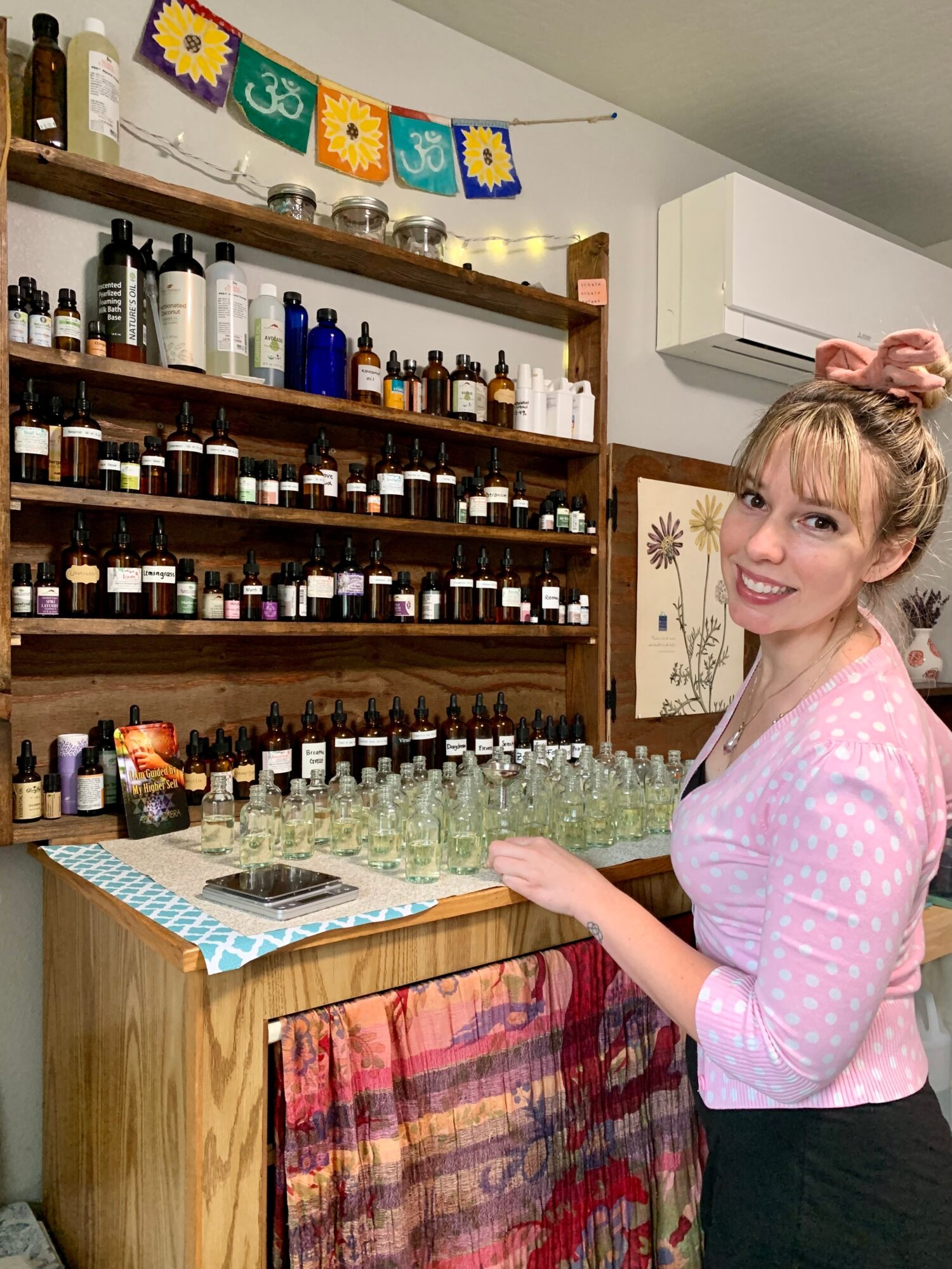 Woman with pink bow in hair, smiling, standing near shelves with bottles and jars, in a room with colorful decorations.