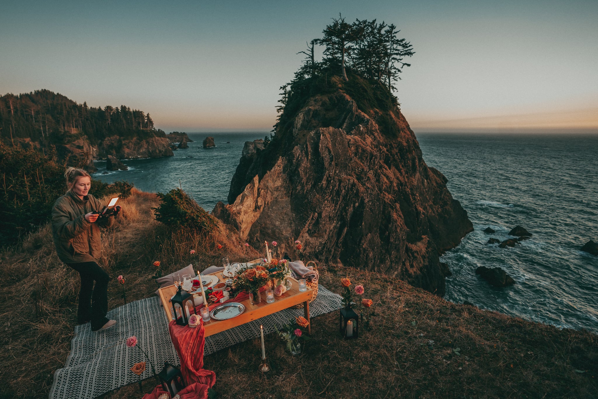 Person standing near a table set for outdoor dining on a cliffside overlooking the ocean, with a large rock formation and trees in background.