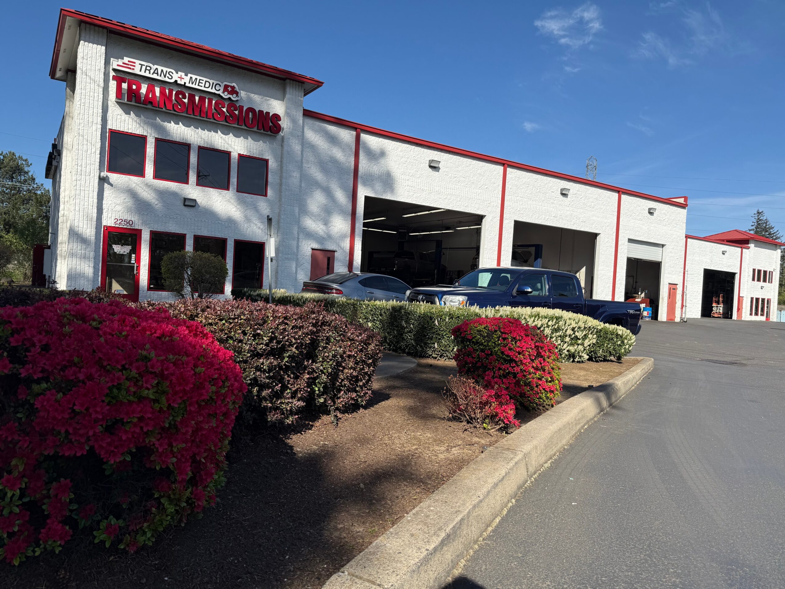 Auto repair shop with a sign reading 'Transmissions' and several cars parked outside, under a blue sky.