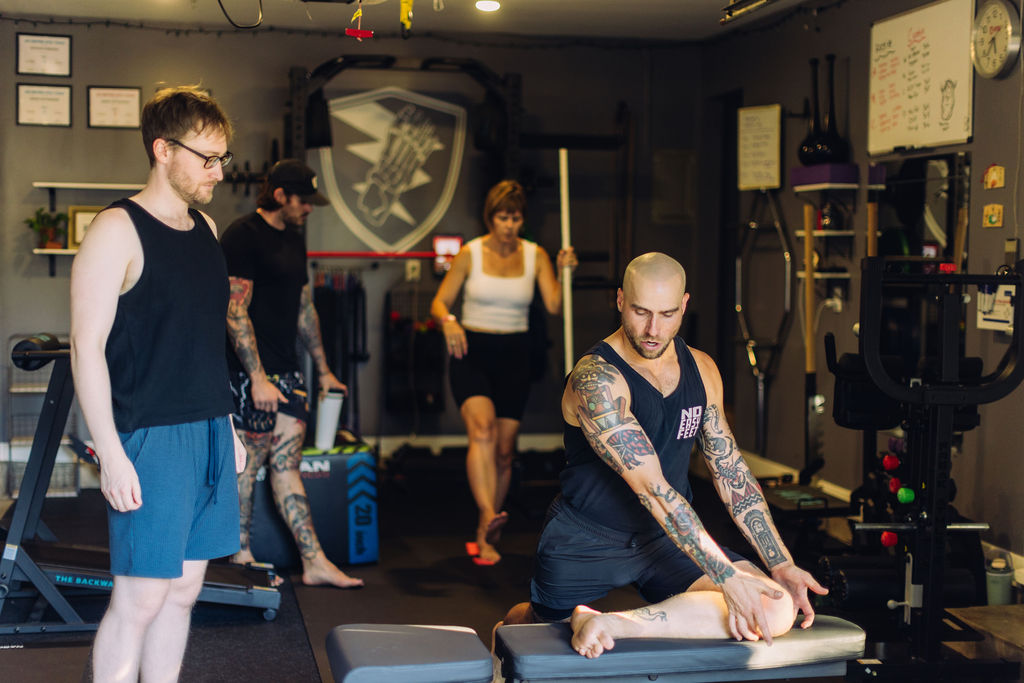 Group of people in a gym, some sitting and others standing, with workout equipment and a logo on the wall.