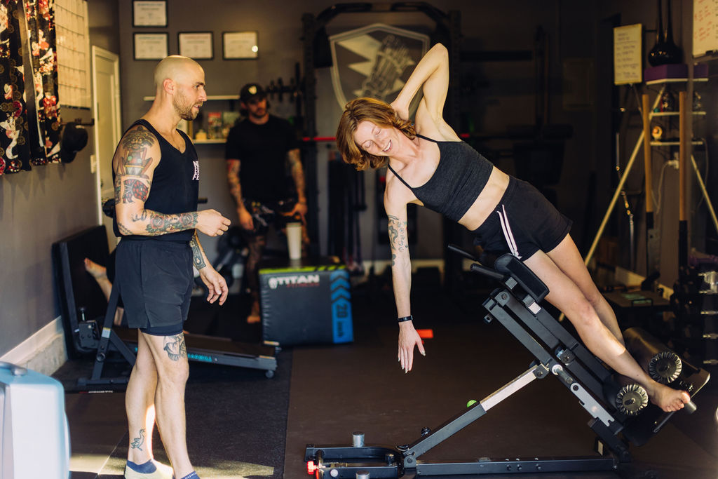 Woman in workout clothes performs a side plank on a bench, with a trainer observing in a gym setting.