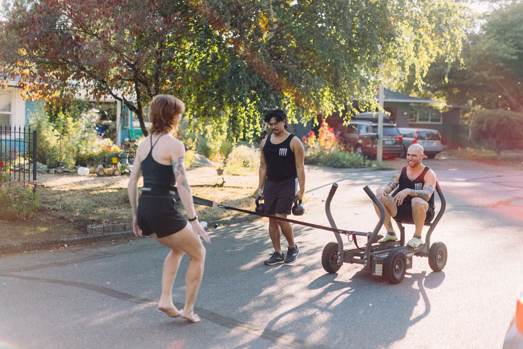 Three people outdoors near trees and a street, one sitting on a cart, two standing, with sunlight filtering through leaves.