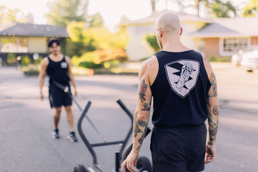 Bald man with tattoos wearing a sleeveless shirt walking outdoors with a person in the background, blurred, sunny day.