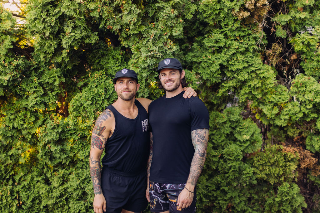 Two men stand close together in front of green foliage, smiling, with one arm around each other's shoulders.
