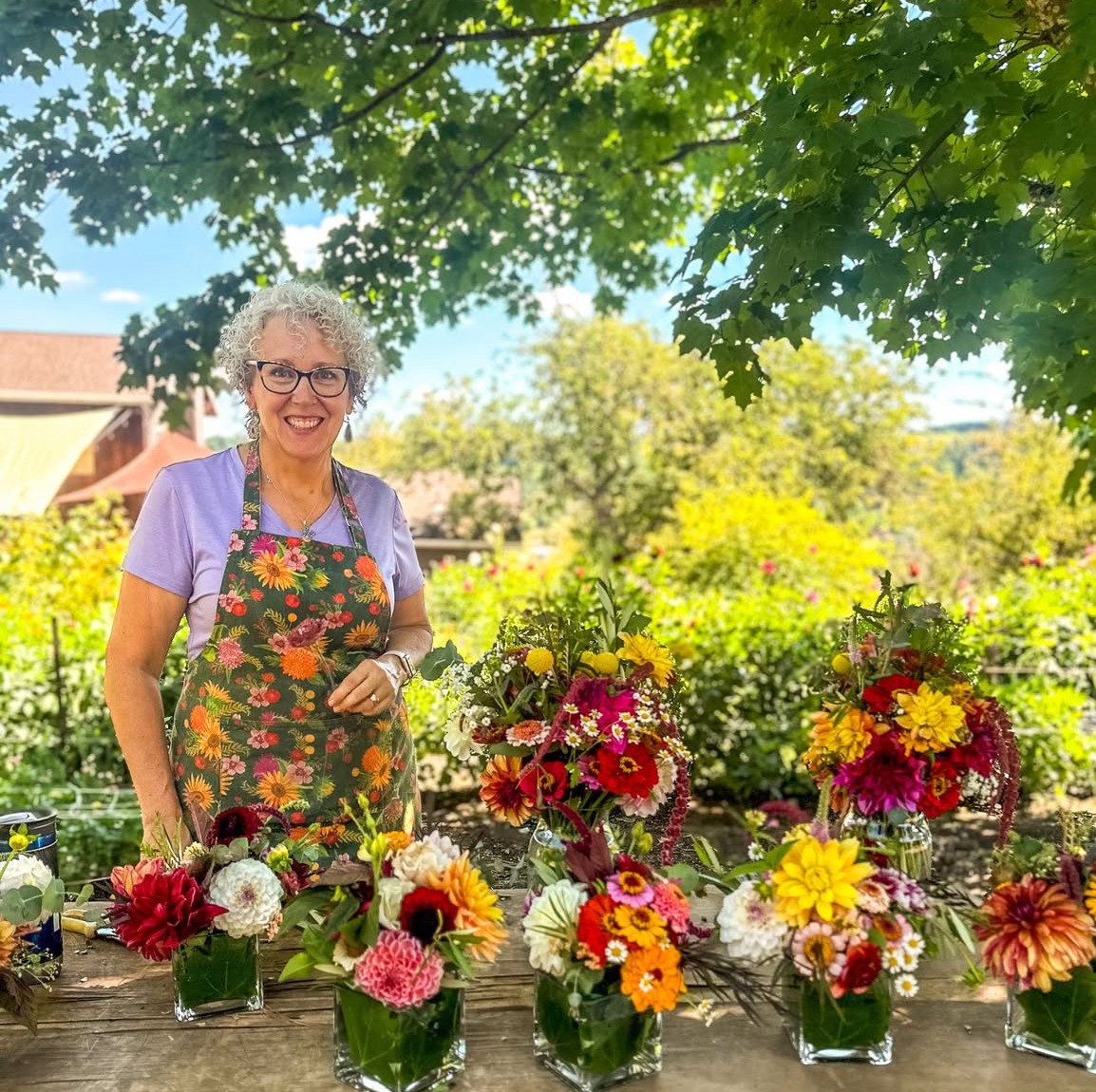 Smiling woman with curly gray hair wearing glasses, purple shirt, and floral apron, standing outdoors with flower arrangements on a table.