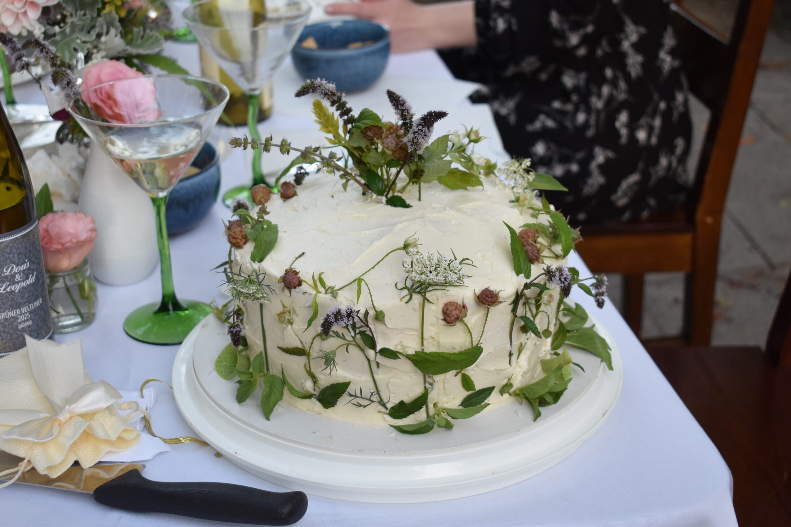 Decorative white cake with flowers and greenery on a table, surrounded by glasses and tableware, with a person in the background.