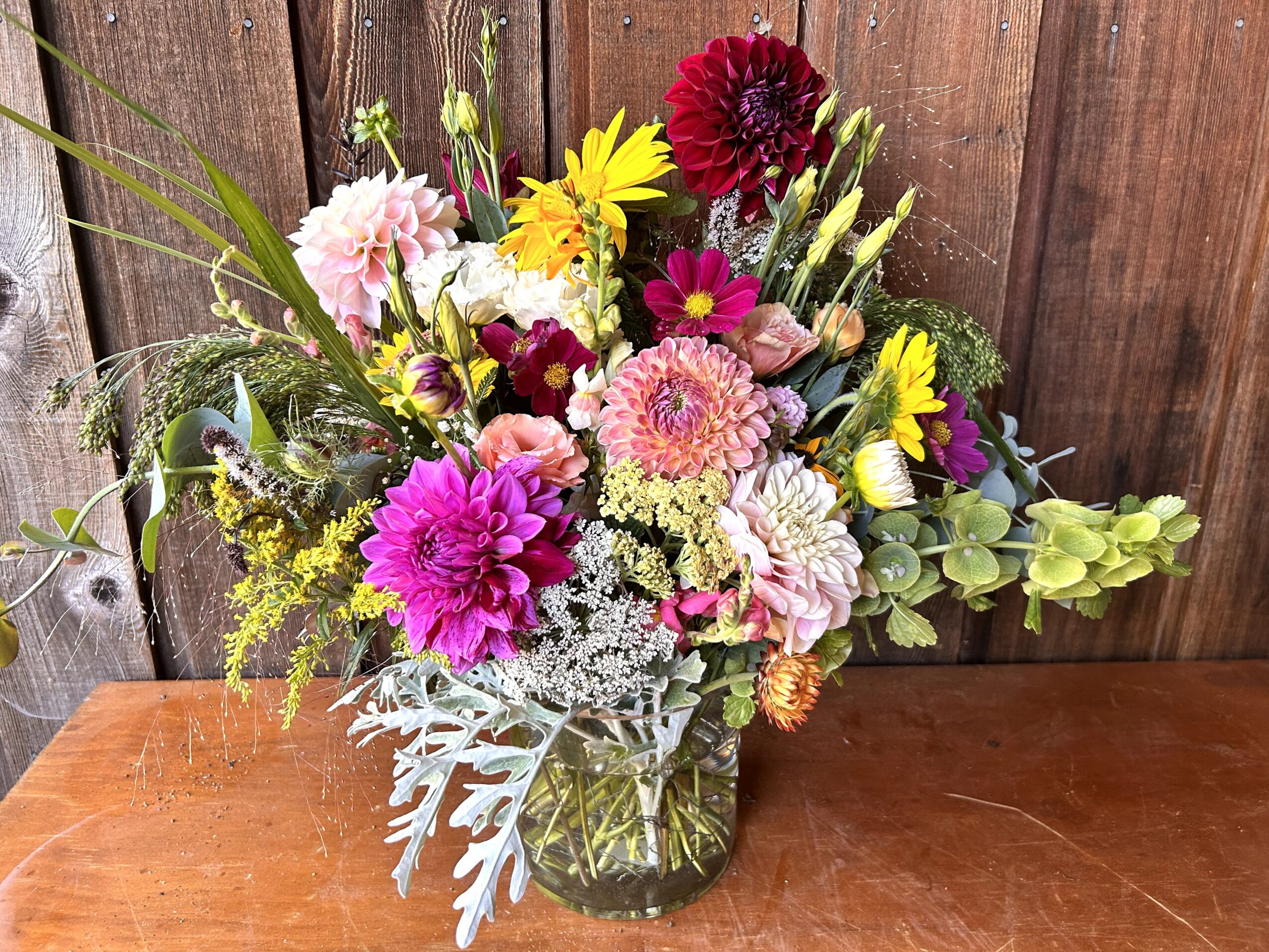 Colorful flower bouquet with pink, yellow, purple, and white flowers in a glass vase on a wooden surface, wooden background.