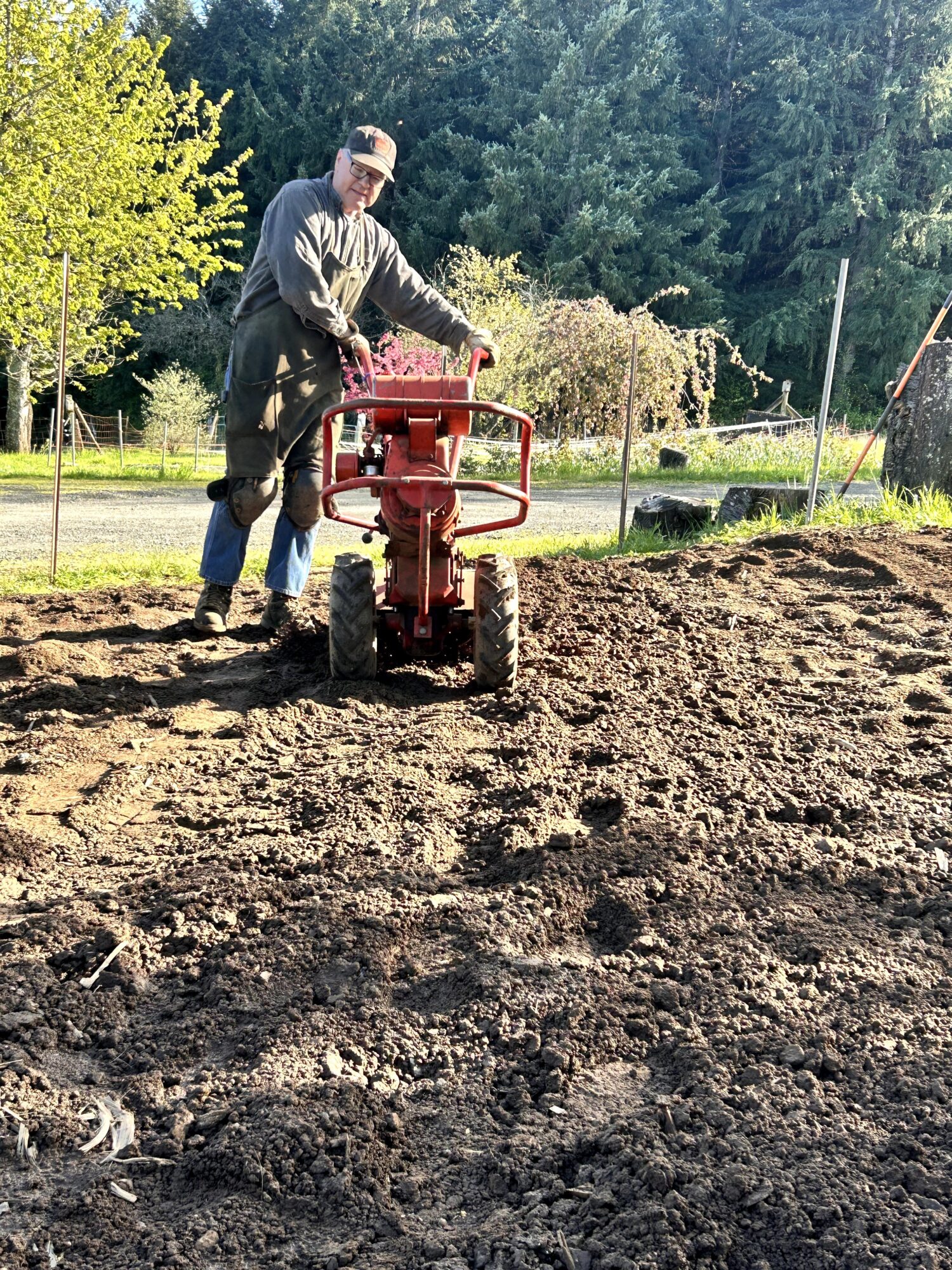 Person operating a red tiller on a dirt field with trees and a road in the background.