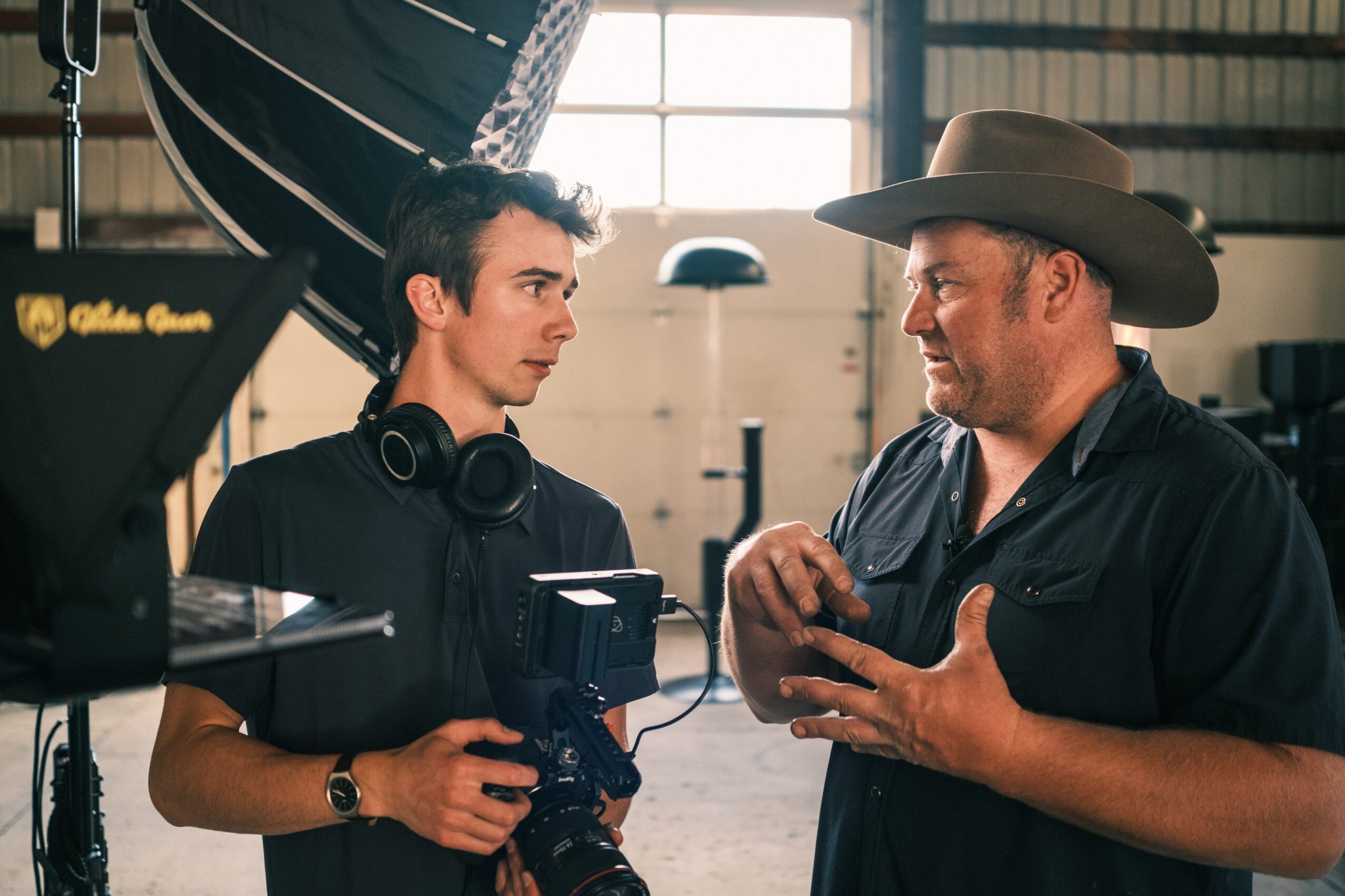 Two men in conversation inside a large industrial space, one holding a camera, the other wearing a cowboy hat, both engaged in discussion.