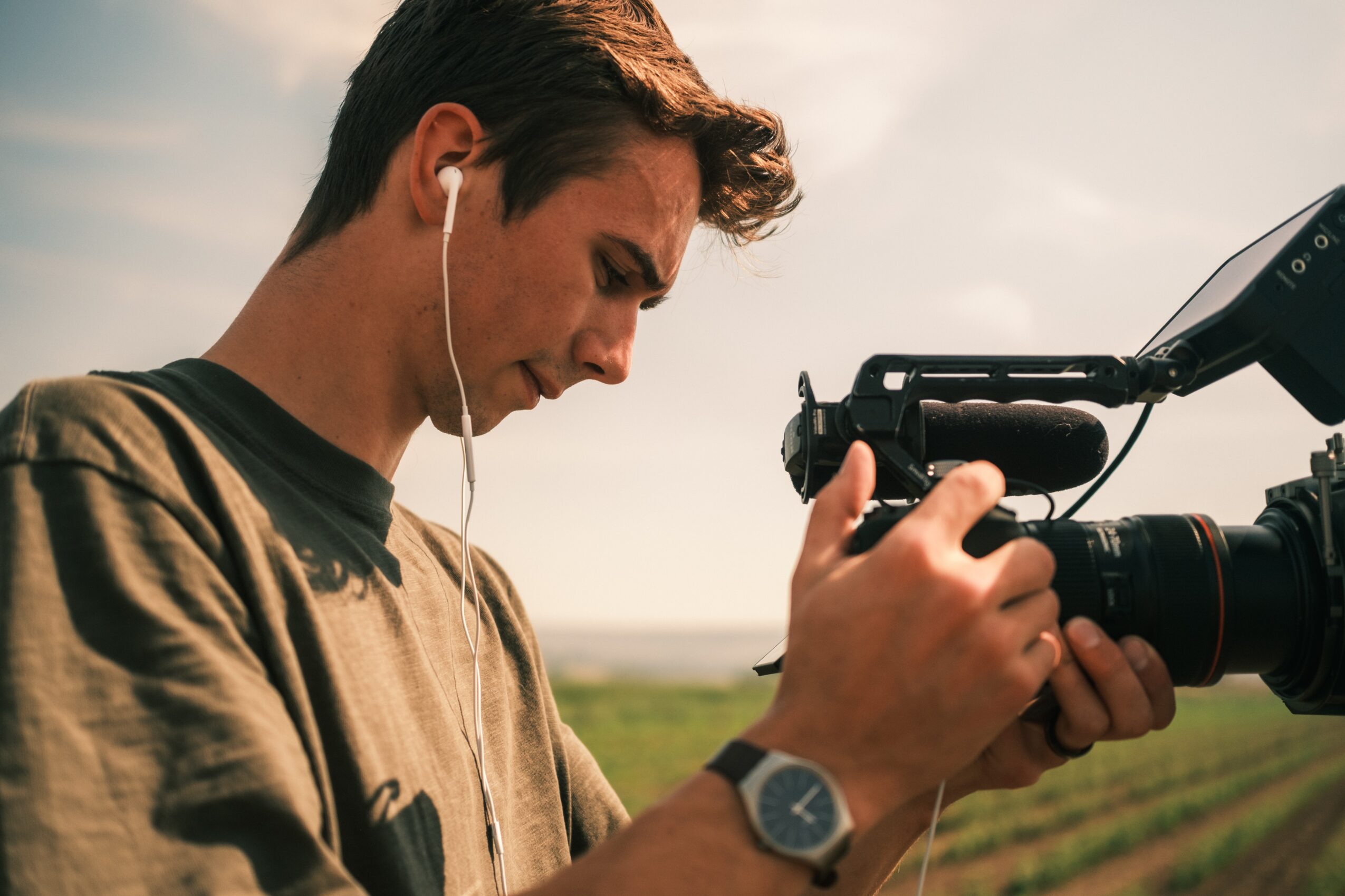 Young man operating a professional camera outdoors, wearing earphones and a watch, with a landscape background.