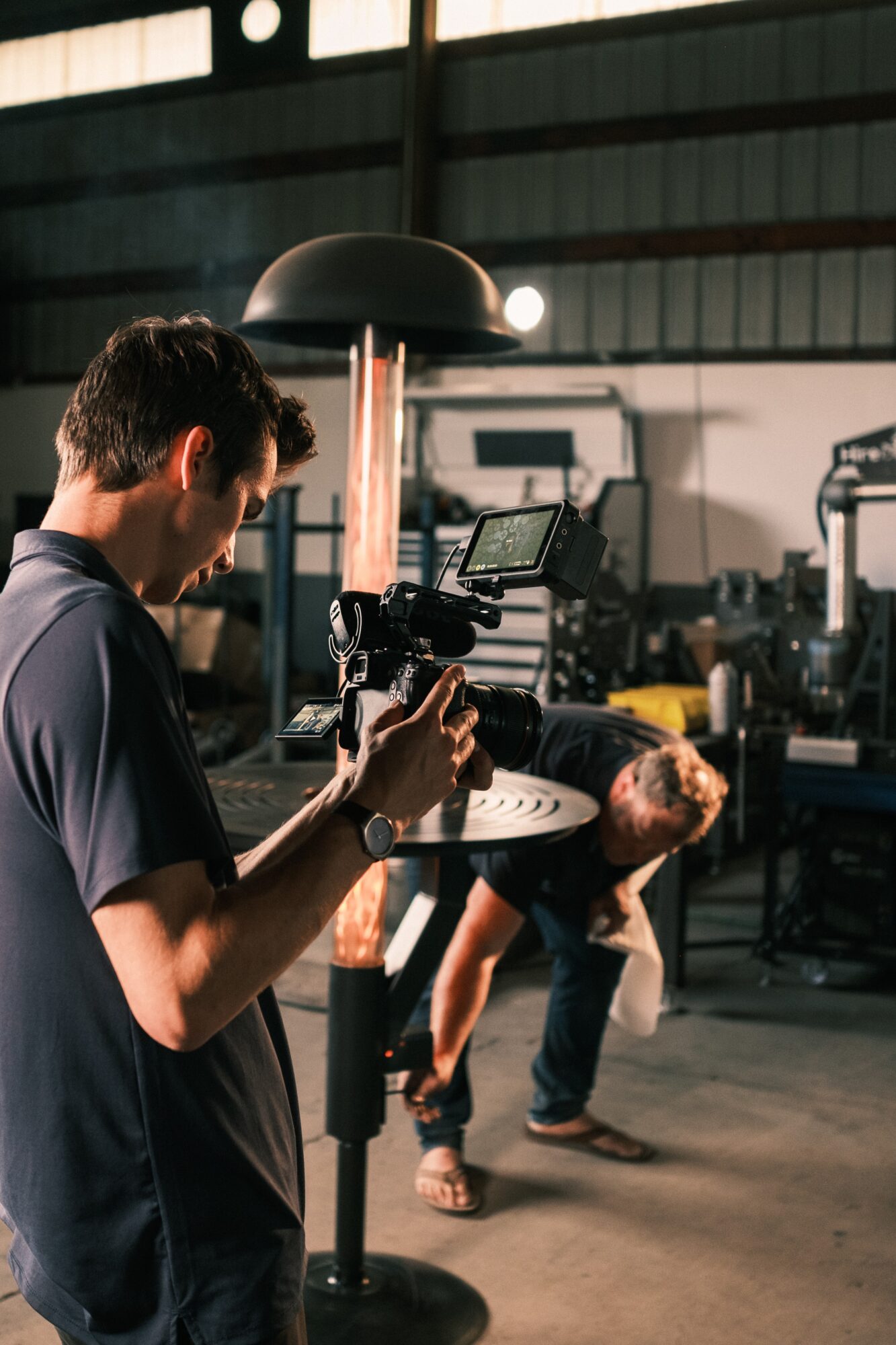 Person operating camera equipment in a workshop with another person working nearby, industrial setting, dim lighting.