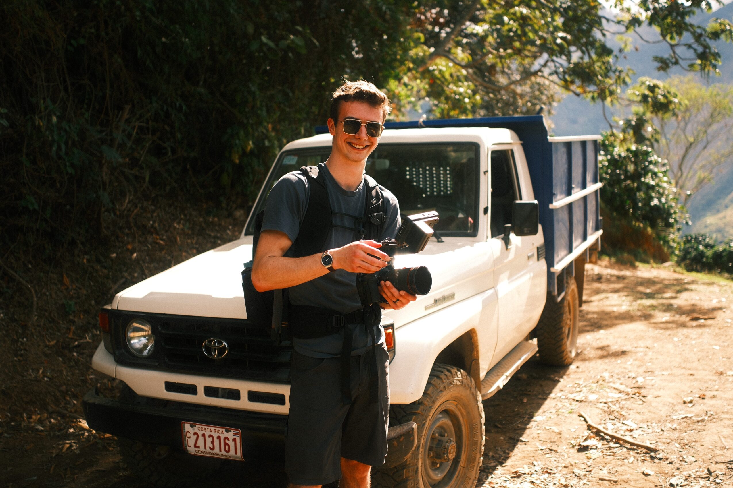 Young man with sunglasses holding a camera in front of a white pickup truck on a dirt path, trees in background.