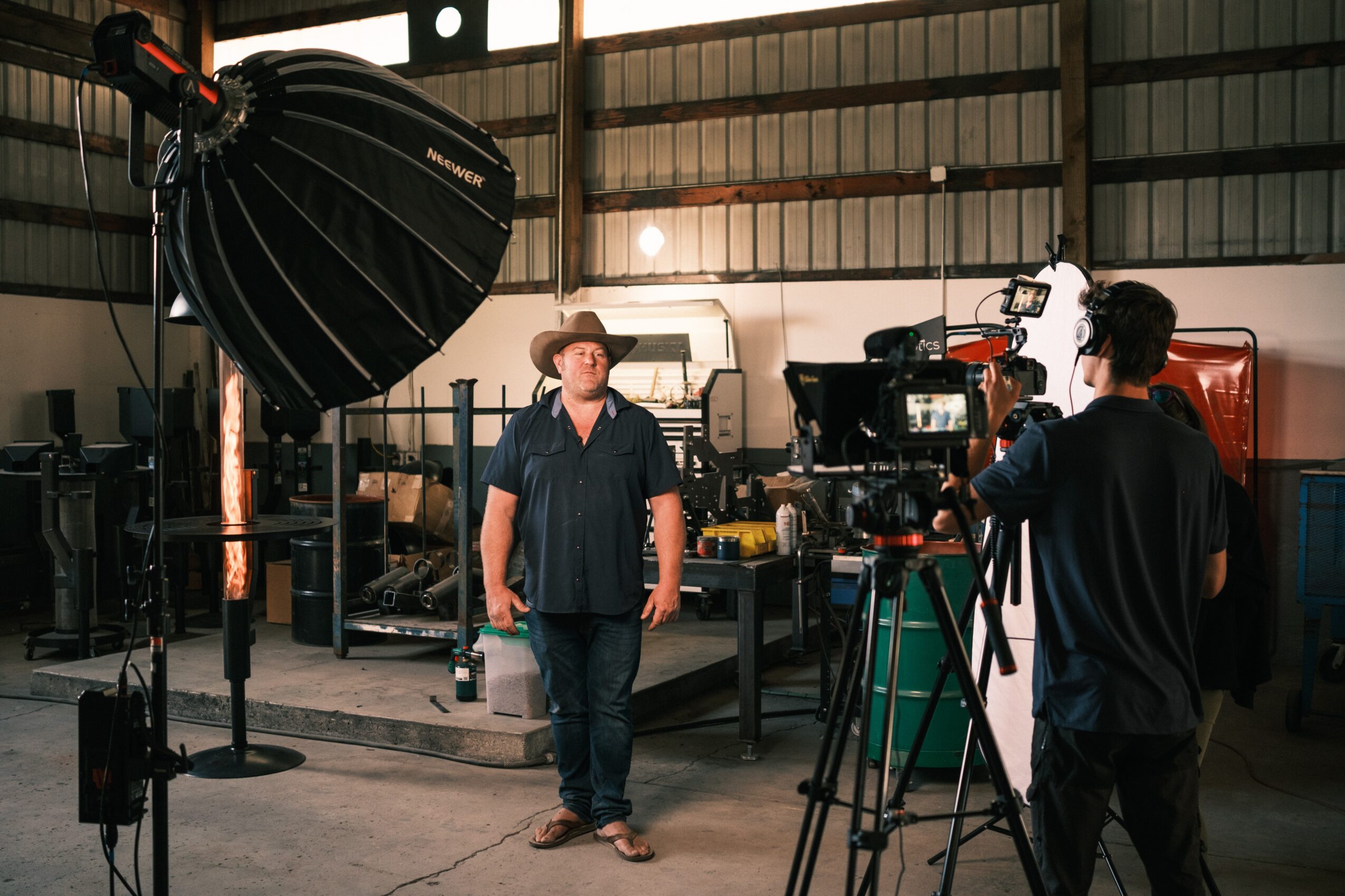Man in cowboy hat and dark shirt standing in industrial workshop with camera crew filming him.