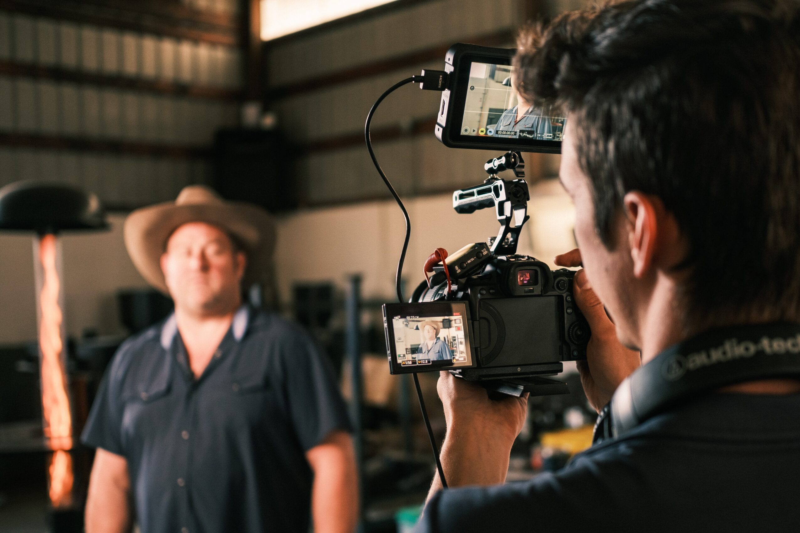 Person filming a smiling man wearing a hat indoors with equipment and industrial background, camera focused on the man.