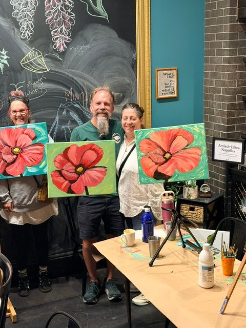 Three people stand behind a table with painting supplies, holding paintings of pink flowers with yellow centers.